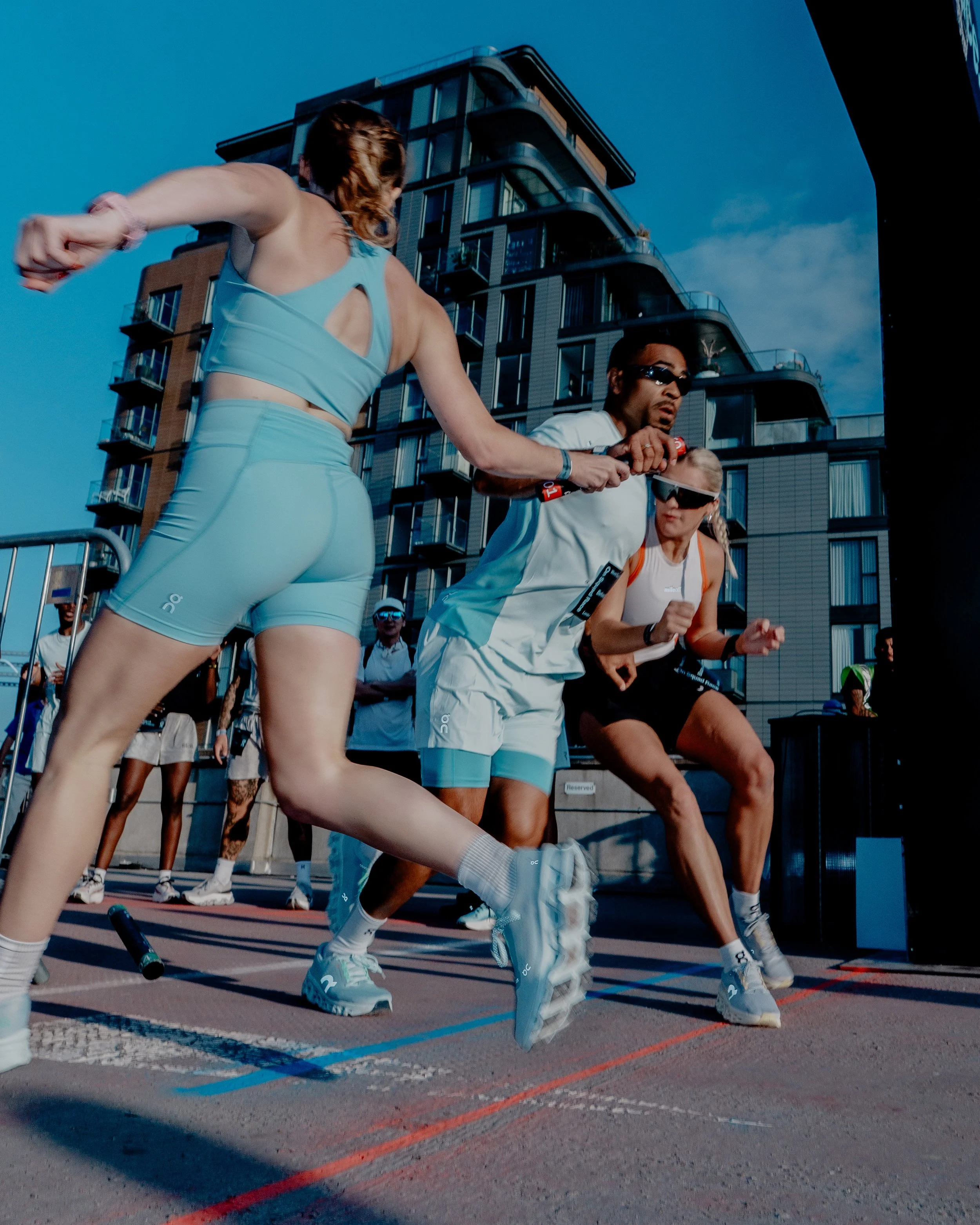 People are participating in an outdoor fitness or martial arts class, performing a move involving knee lifts or kicks, with a modern building and a clear blue sky in the background.