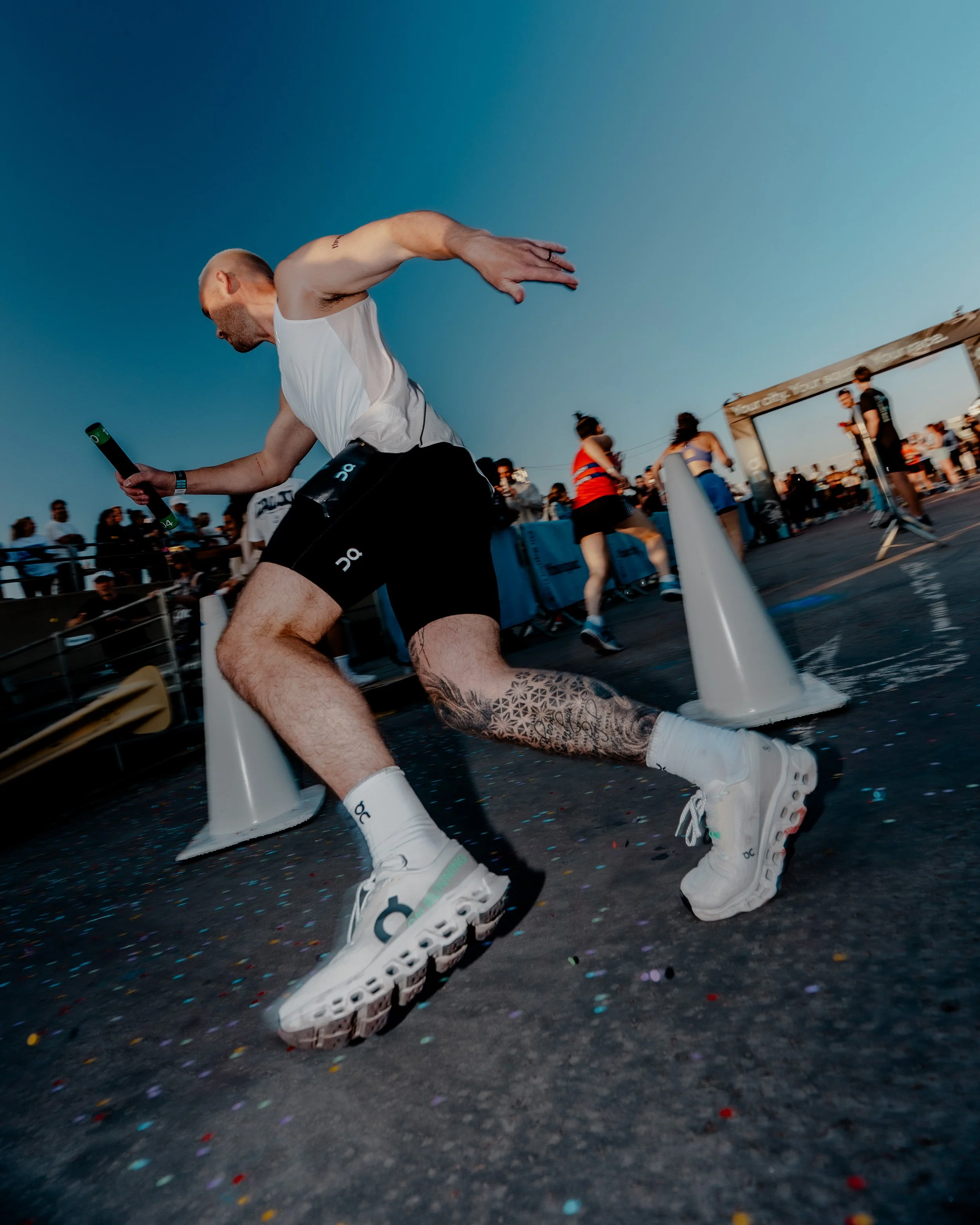 A man running in a marathon or race wearing a white tank top, black shorts, and white sports shoes. He is holding a green object in his right hand and has a tattoo on his lower left leg. Other runners and spectators are visible in the background, wit
