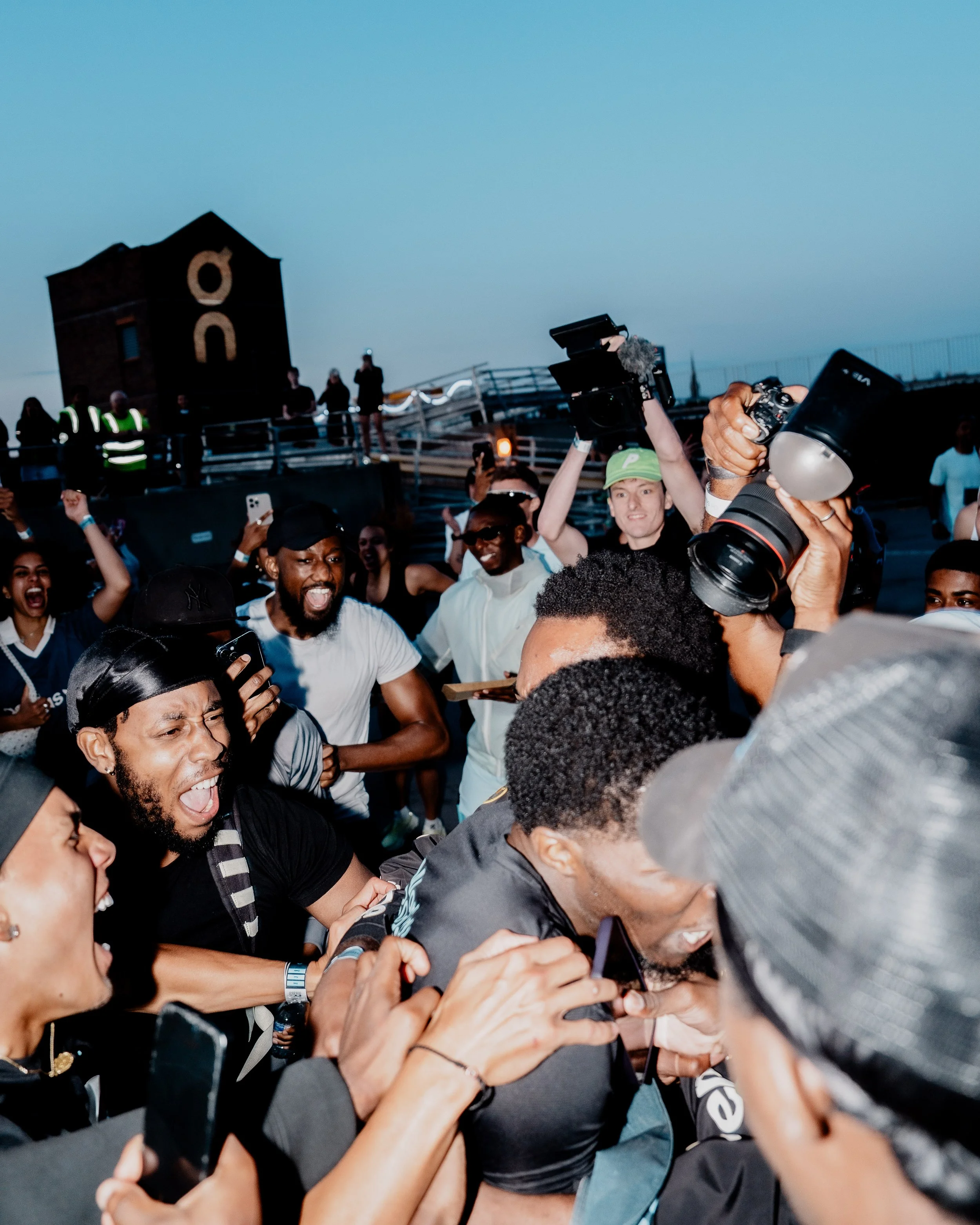 A crowd of people gathered together, some smiling, with many holding up smartphones and cameras, in front of a structure with a black building and white symbols in the background.