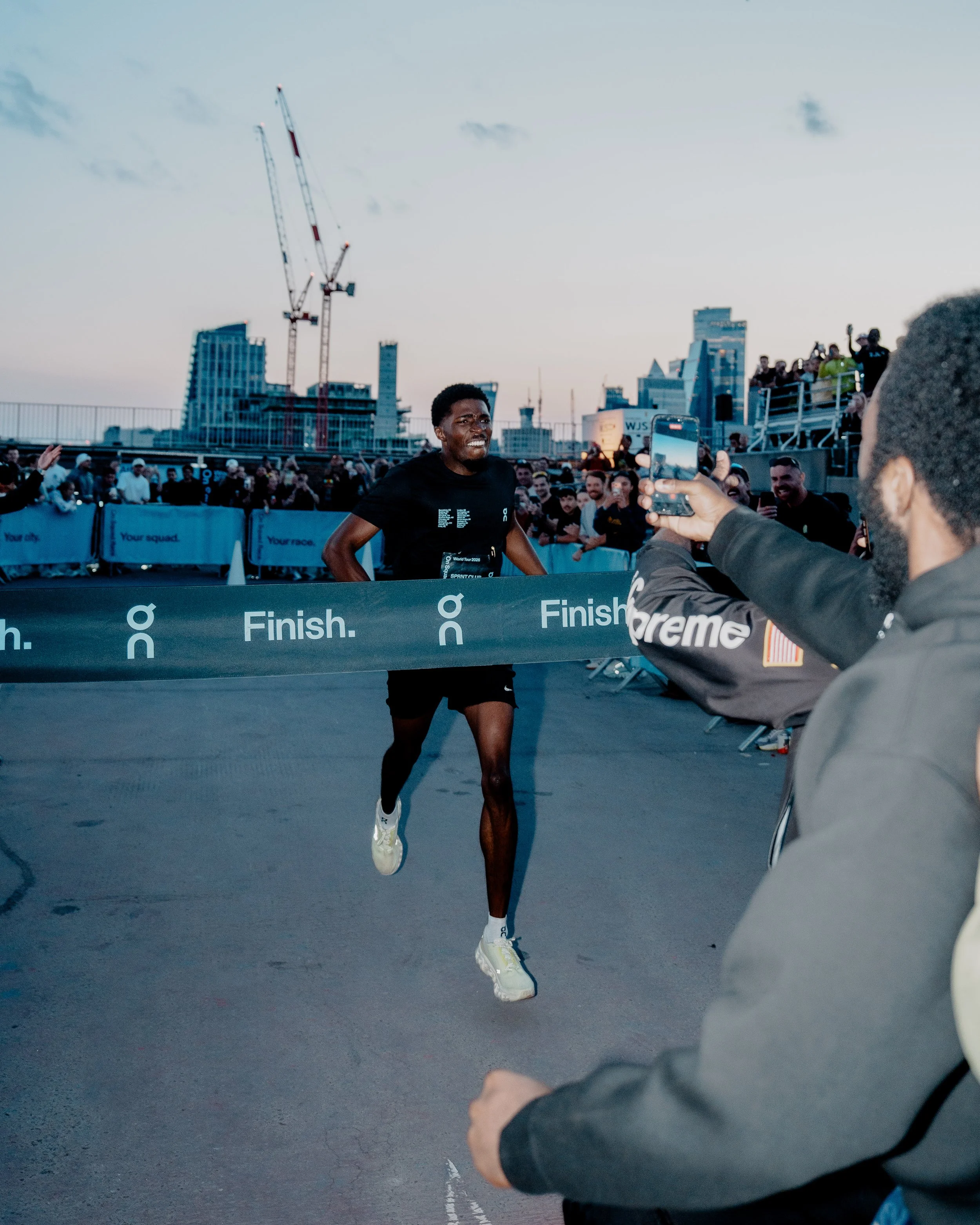 A male runner crossing the finish line in a race with a city skyline in the background, a crowd cheering, and a person taking a photo.