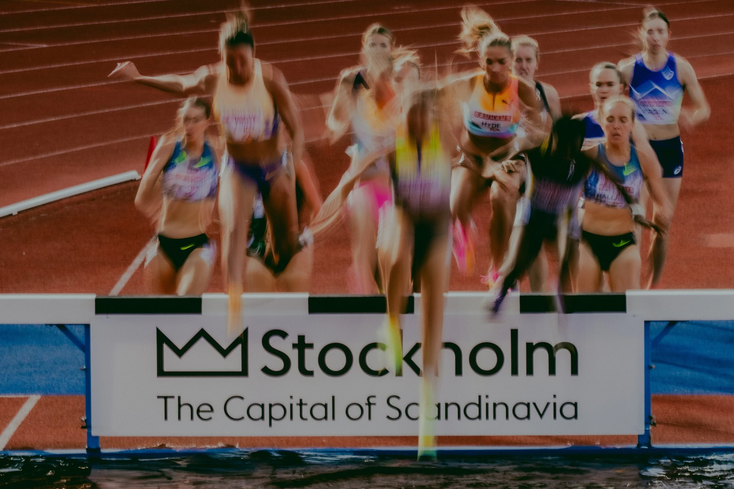 Several female athletes running in a race on a red track at a stadium, with a banner in front that reads "Stockholm, The Capital of Scandinavia".