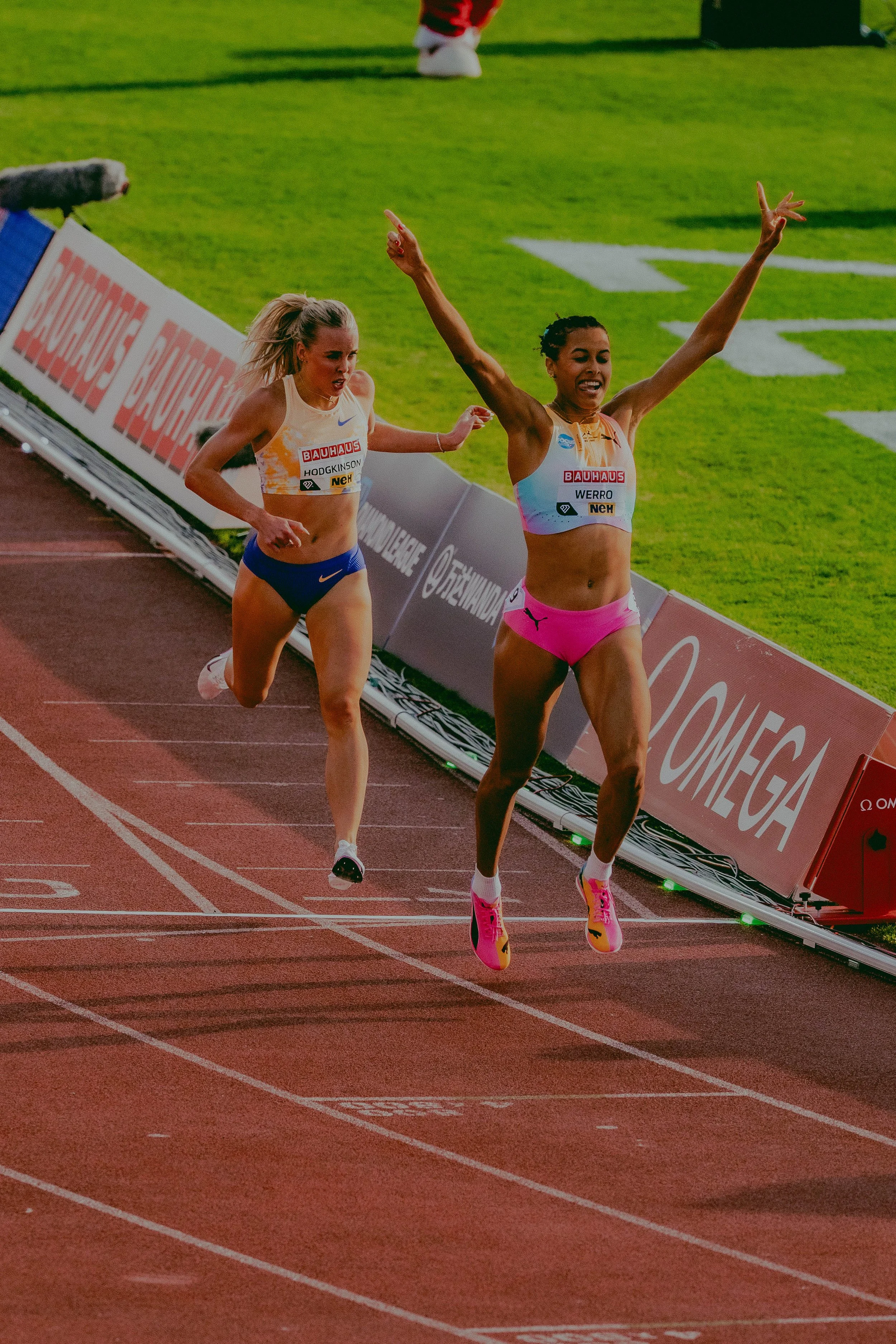 Two female athletes crossing the finish line in a track race, with one celebrating and the other appearing exhausted. They are on a running track with green grass and advertising banners on the side.