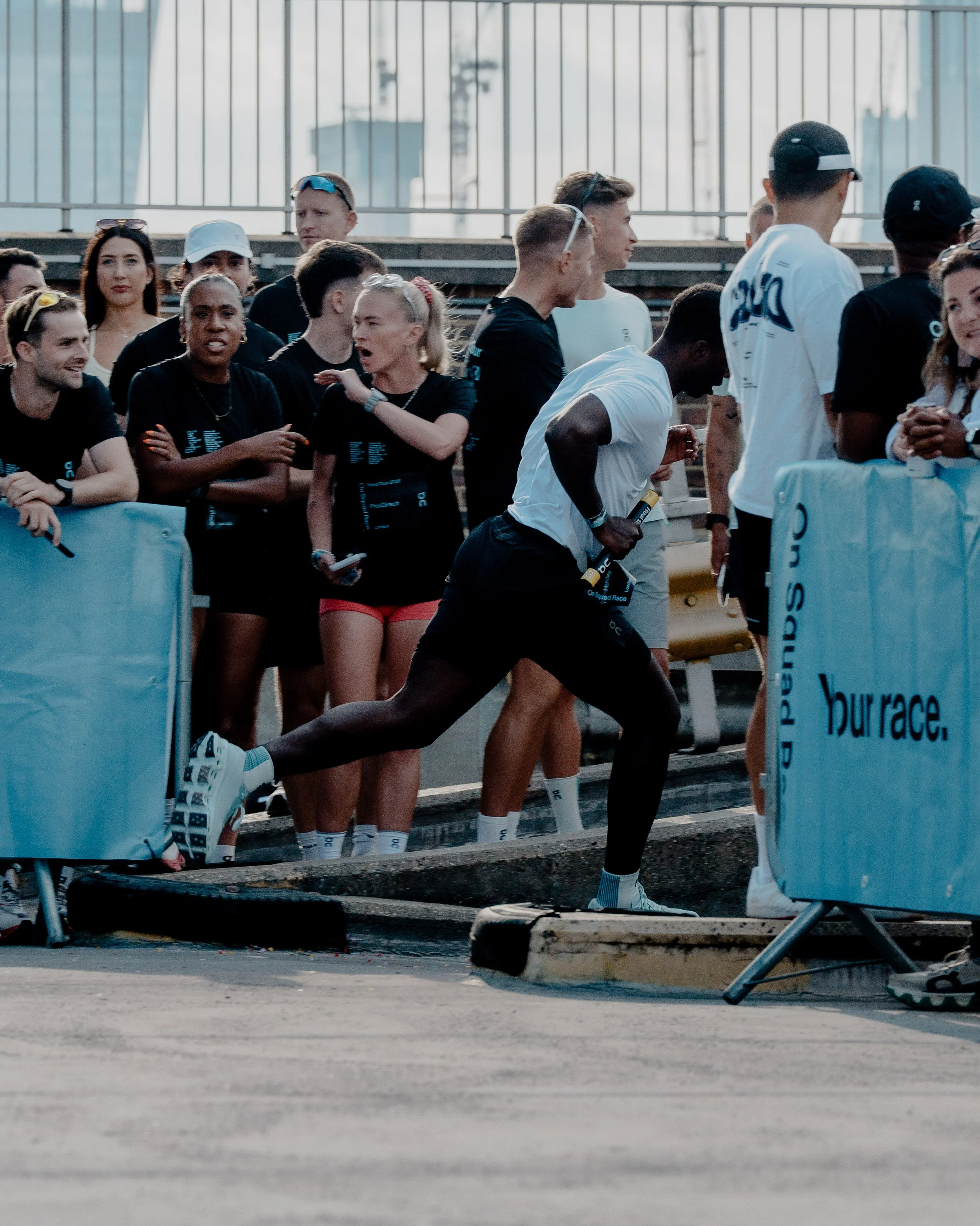 Group of athletes preparing for a race at the starting line, with some stretching, and others engaging in conversation, on a track with barriers and a cityscape in the background.