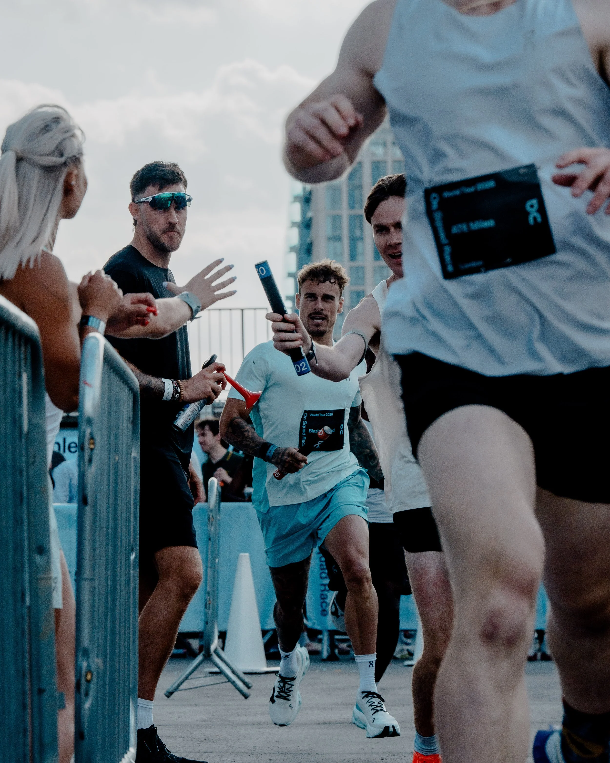 A group of runners participating in a race, with some runners passing through a designated area marked with barriers. One runner in the center is holding a baton, suggesting it might be a relay race. The scene is set outdoors with buildings and a clo