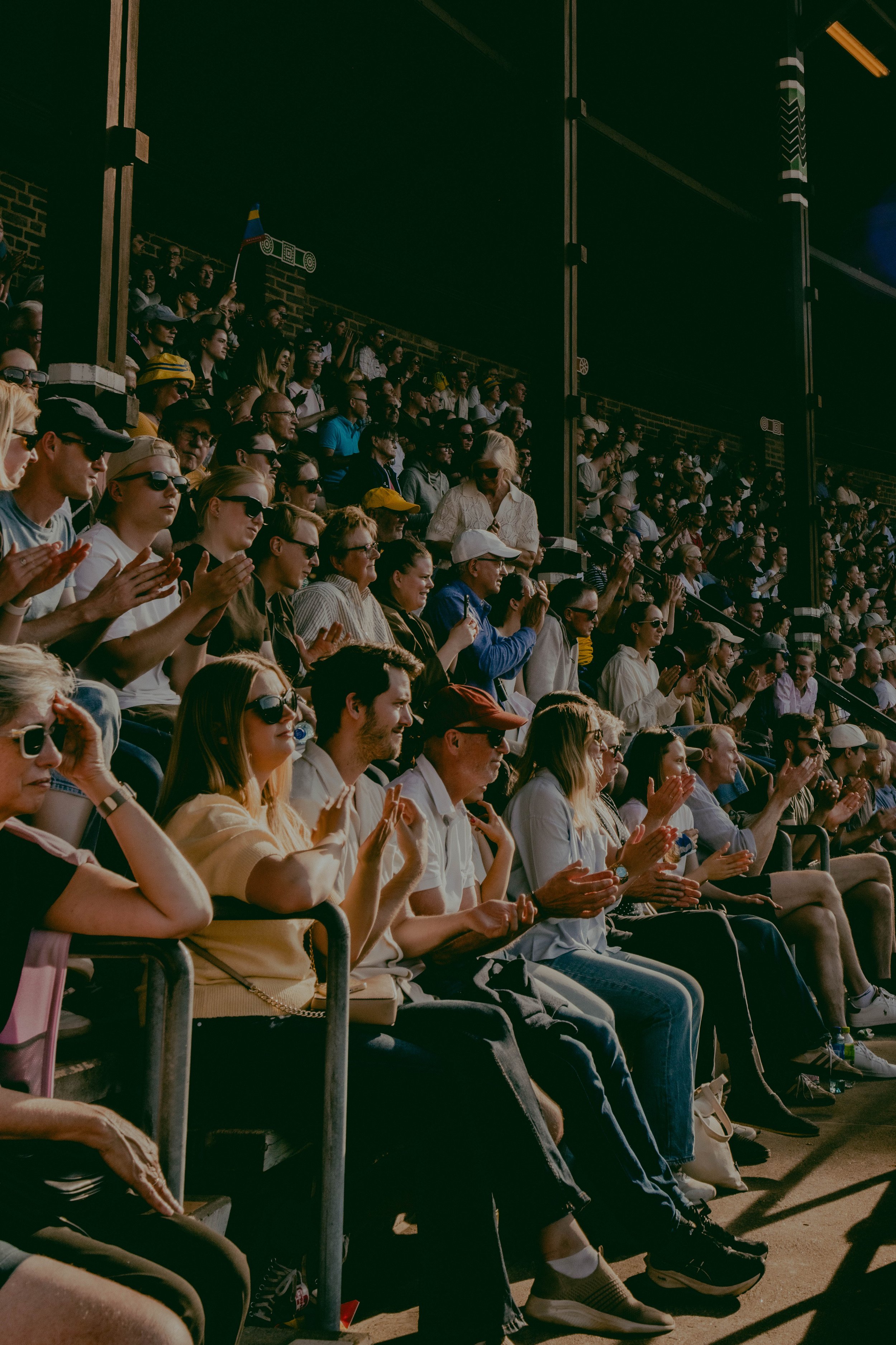 A large crowd of people sitting in stadium seats, watching an outdoor event, with some clapping and others wearing sunglasses and casual clothing.