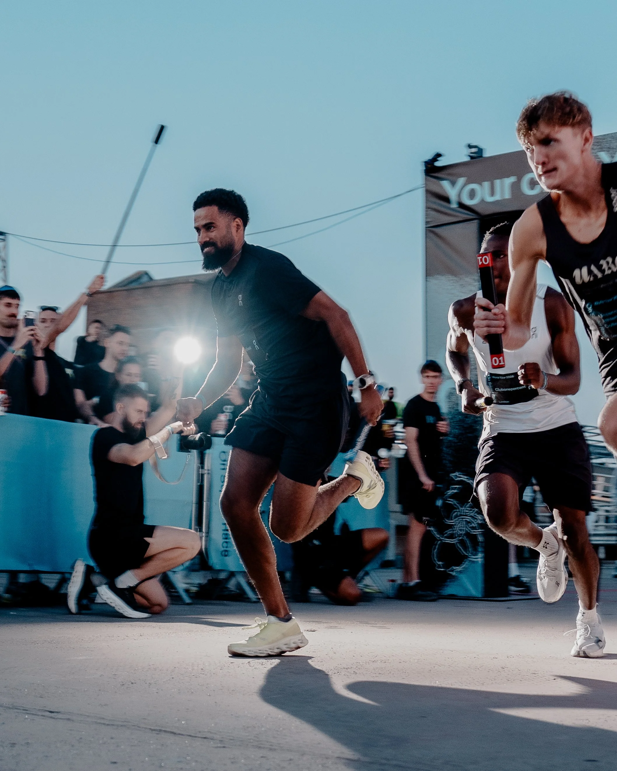 Two runners competing in a race, one in white and black attire holding a baton, and one in black shorts and shirt, with spectators and event banners in the background during the evening or dusk.