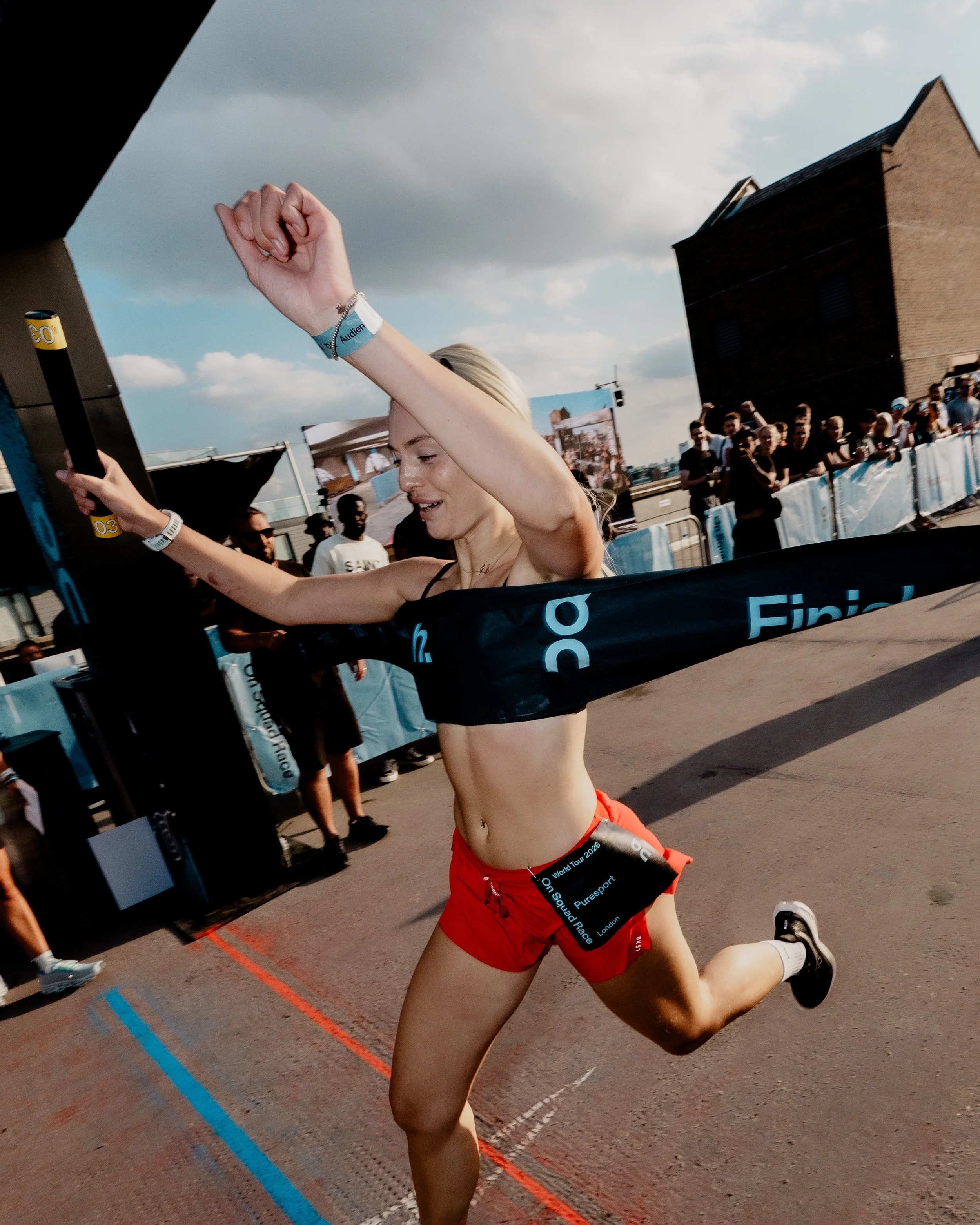 A female runner in red shorts and black sports bra crossing a finish line at an outdoor race event, with spectators and other participants in the background under a partly cloudy sky.