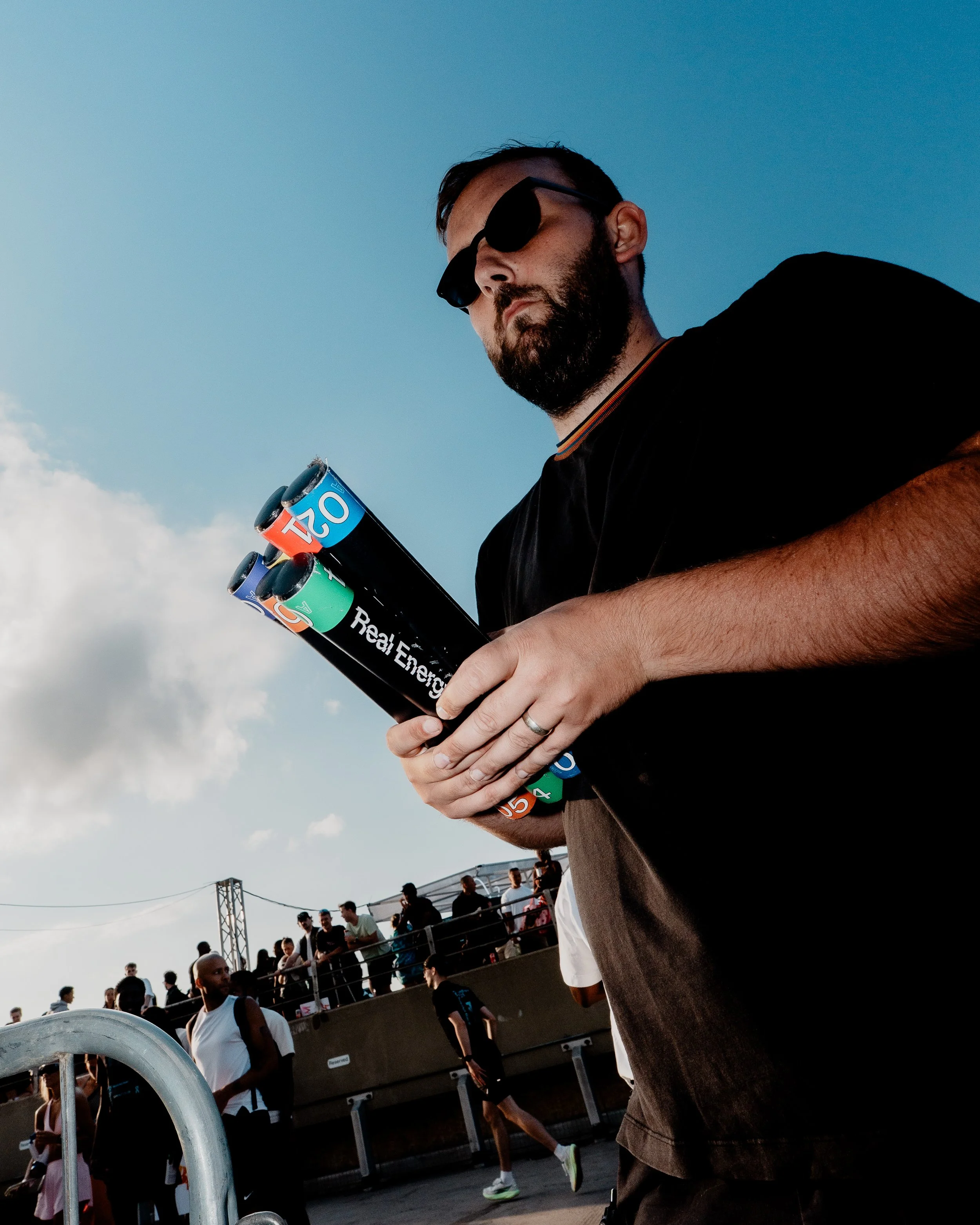 A man with a beard and sunglasses holding a pile of energy drink cans, wearing dark clothing, at an outdoor event with a crowd in the background.