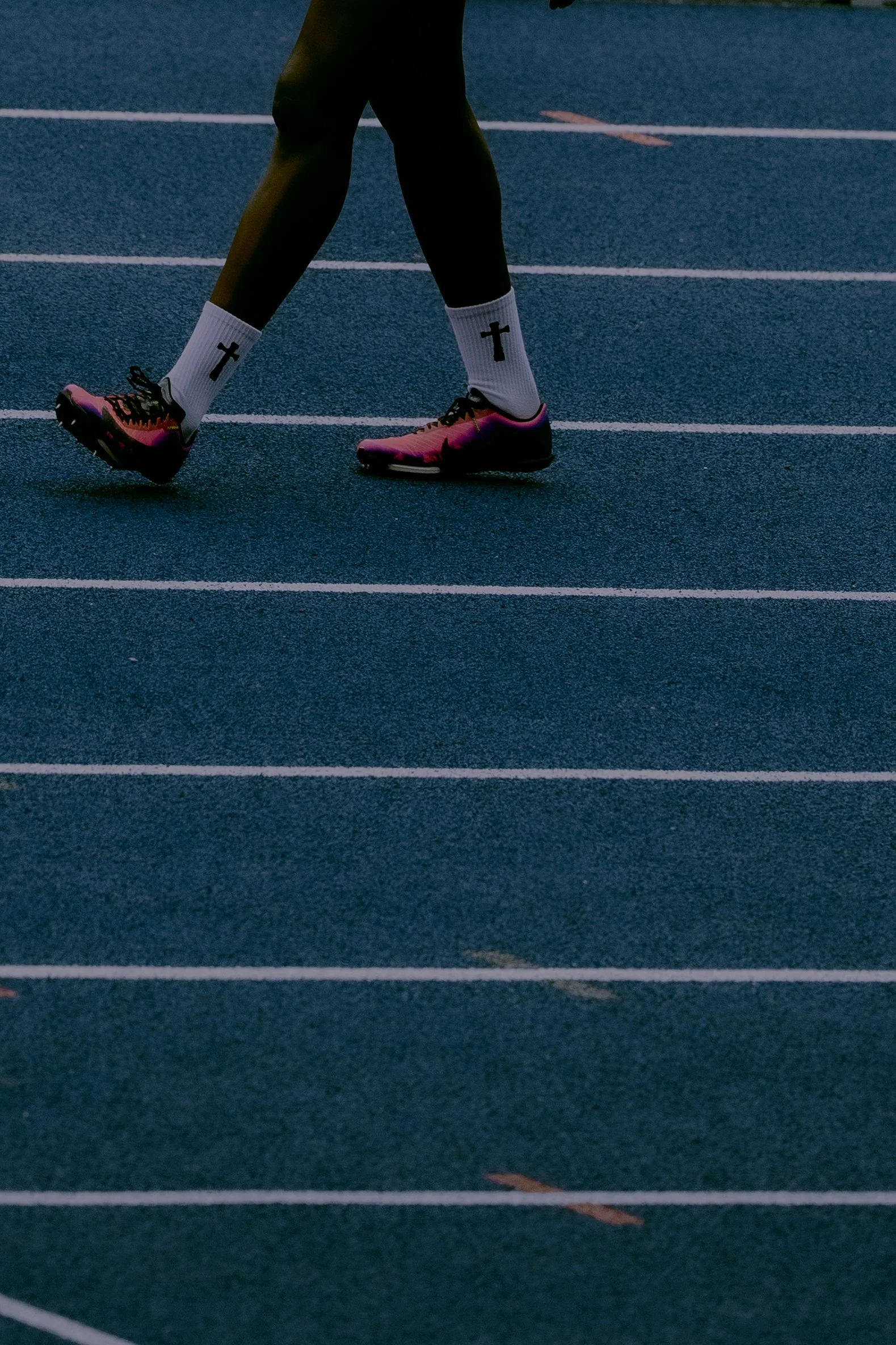 A person walking on a running track wearing white socks with a black cross, pink and black running shoes, and black athletic pants.