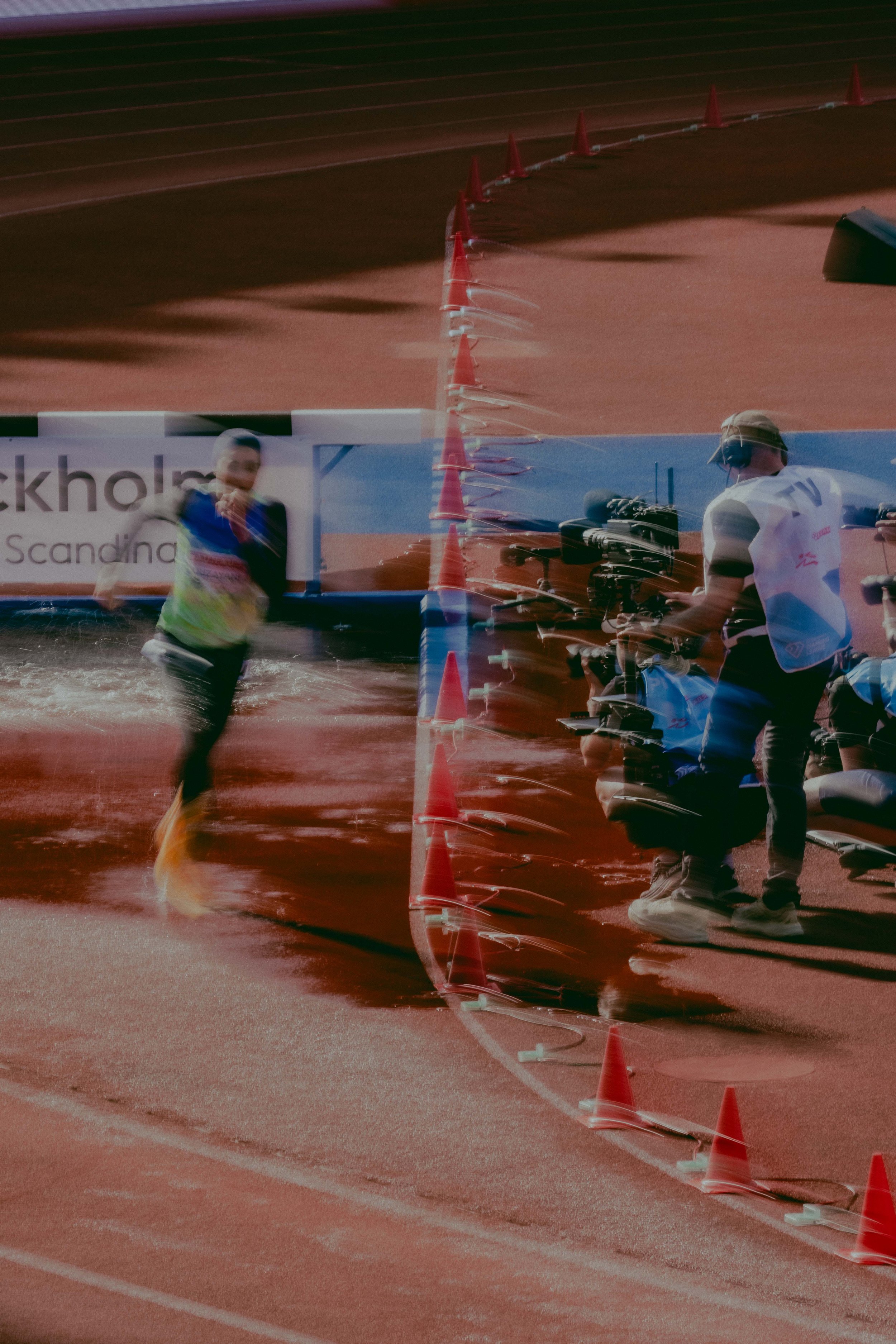 An athlete running on a track near a water obstacle during a race, with a person filming or photographing and a cameraman nearby.