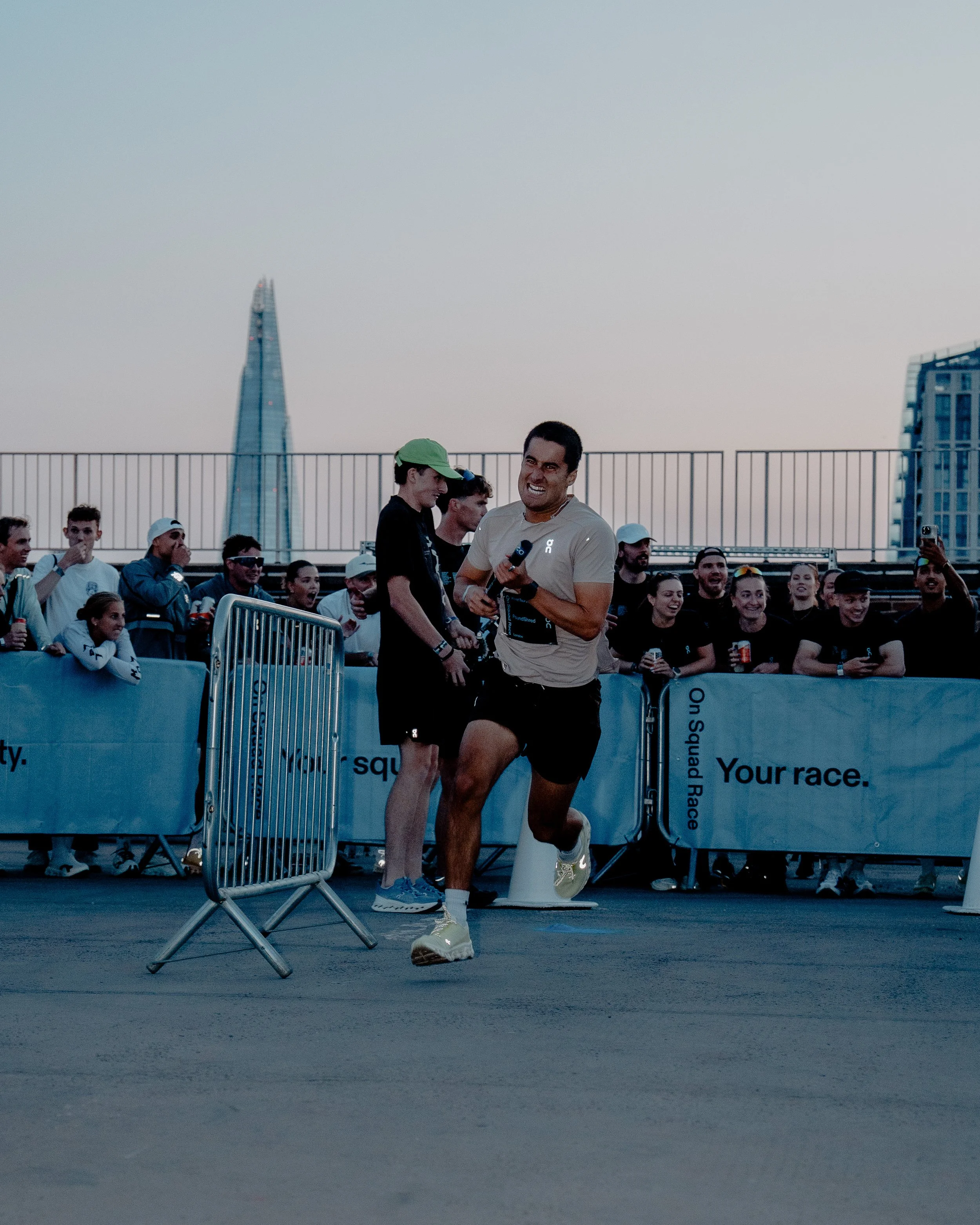 A man in athletic clothing with an eye patch crossing the finish line of a race, smiling. Behind him, a crowd of spectators and supporters cheer. The setting appears to be an urban outdoor event, with high-rise buildings and a clear sky in the backgr
