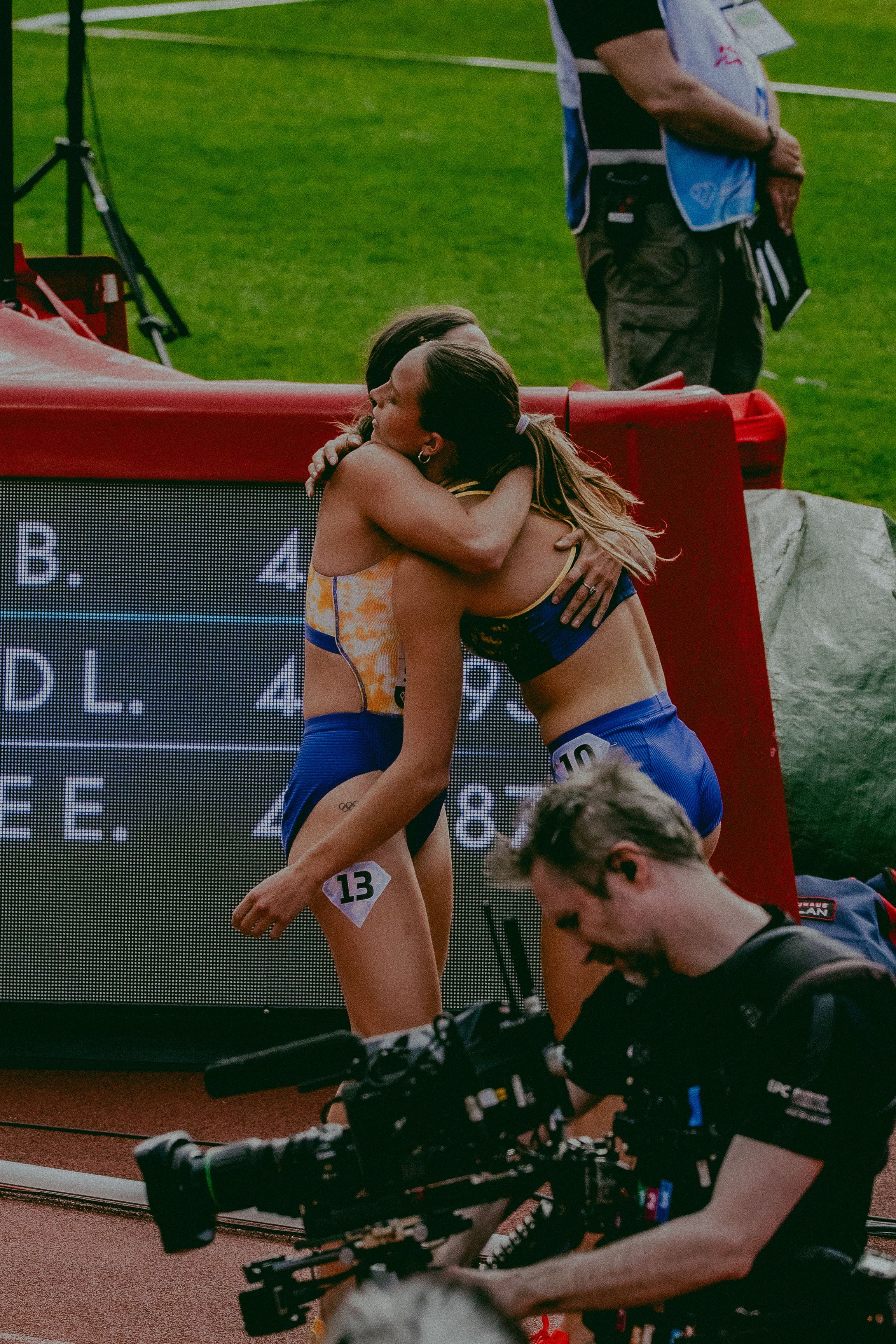 Two female athletes wearing athletic gear hugging each other after a competition on a track field, with a man operating camera equipment in the foreground and a scoreboard in the background.