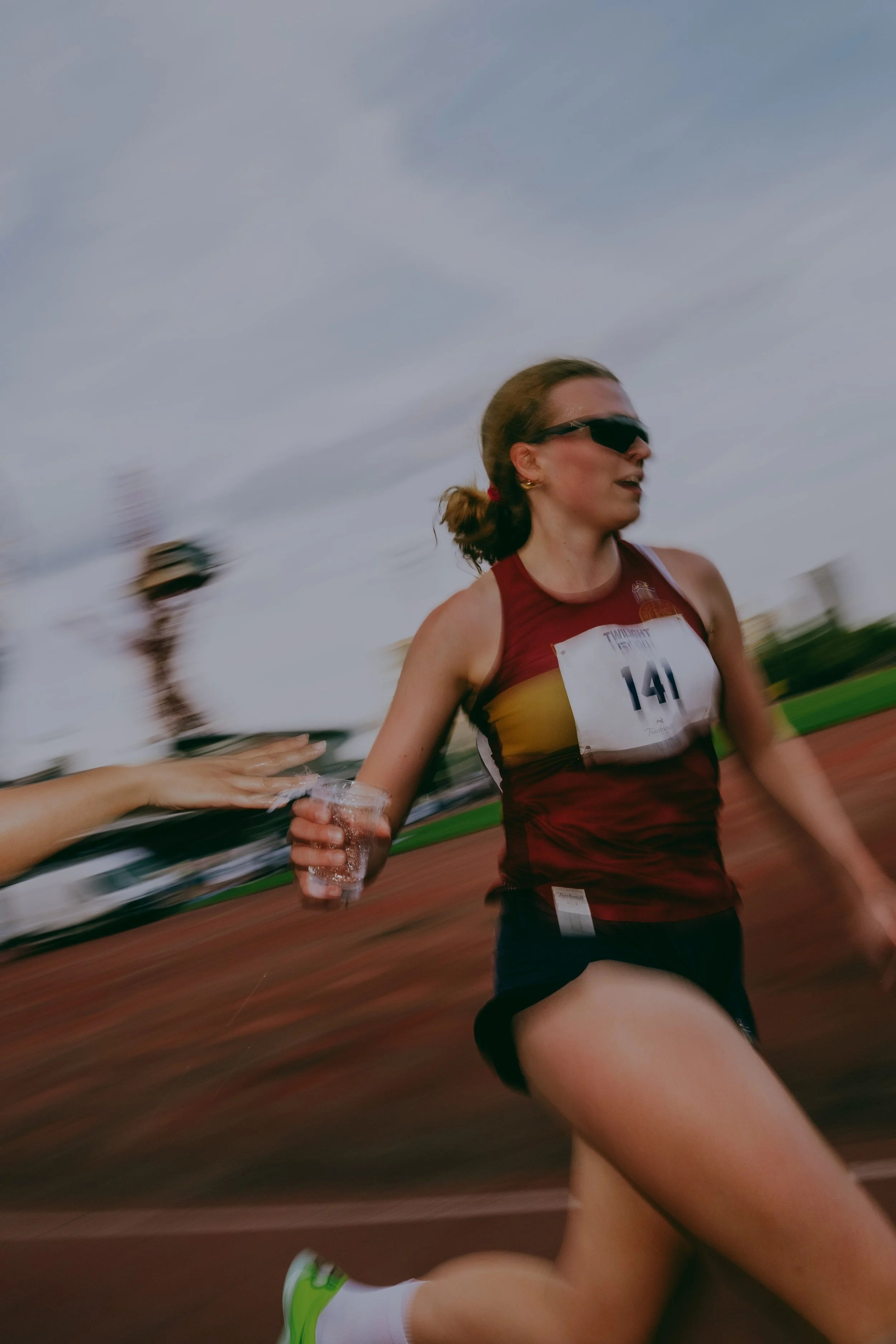Female athlete running in a race, wearing sunglasses, a red sports top, black shorts, and a race bib with the number 141, holding a cup of water, with another runner in the background.