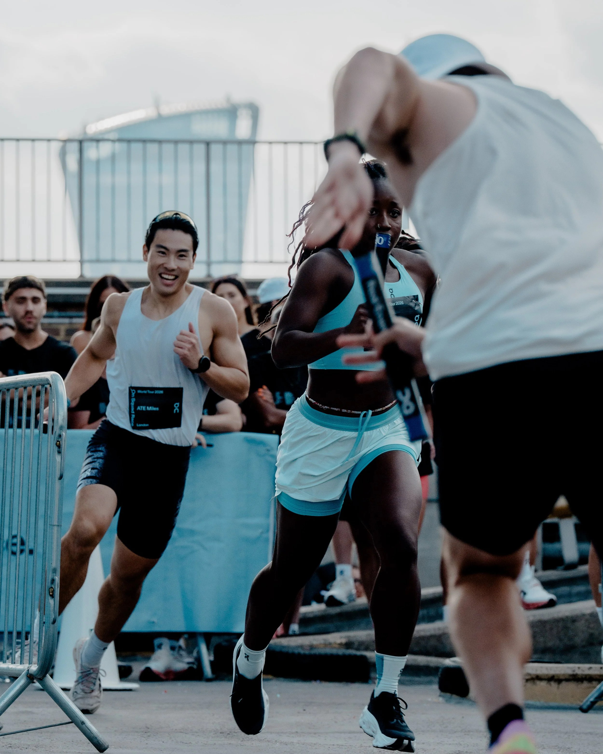 A group of runners participating in a race, with a woman in black running shoes in the foreground, a man with dark hair smiling in a white tank top, and several other runners behind them, all near a barrier on a city street with a modern building vis