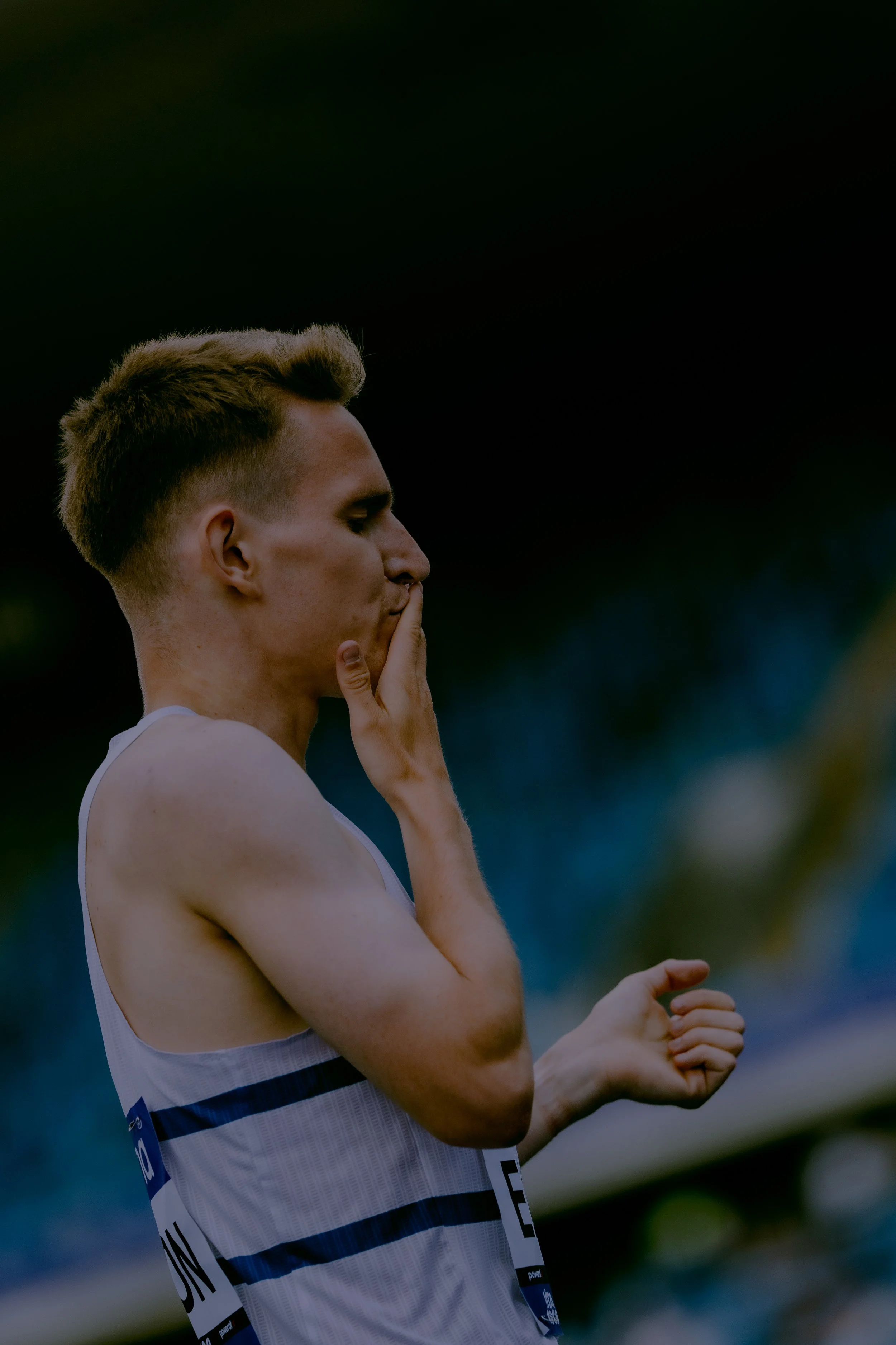 A male athlete wearing a white sports tank top, holding his hand up to his face in a thoughtful pose during a sports event.