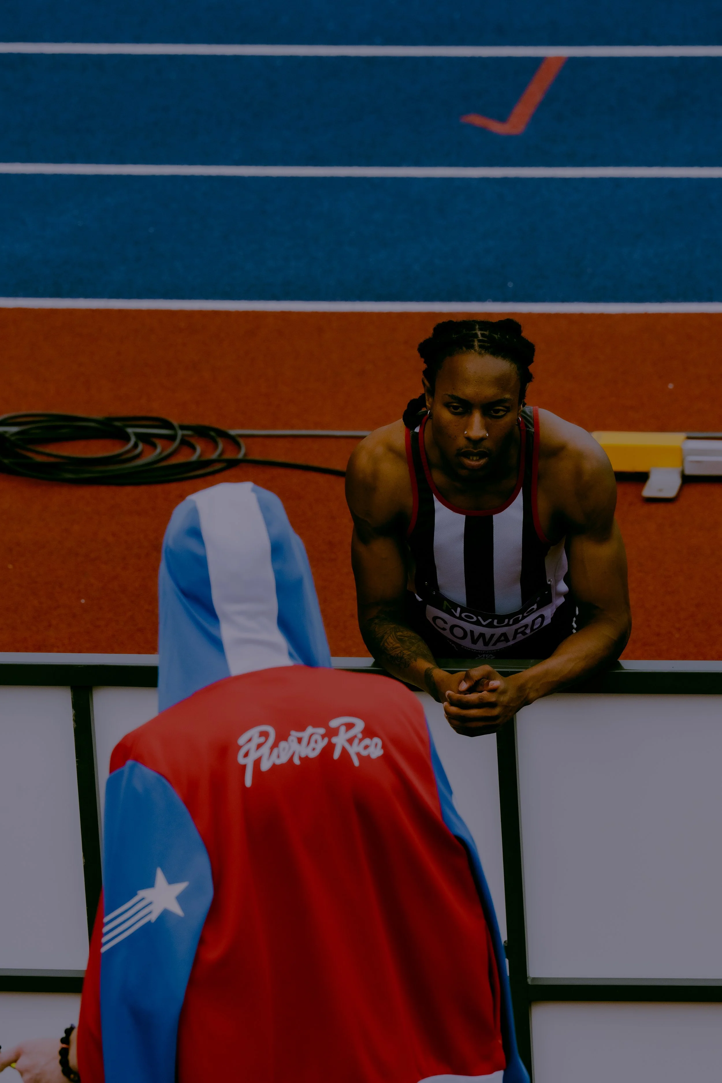 An athlete wearing a red jacket with the words 'Puerto Rico' on the back in front of a woman with braided hair, on a track with lanes marked.
