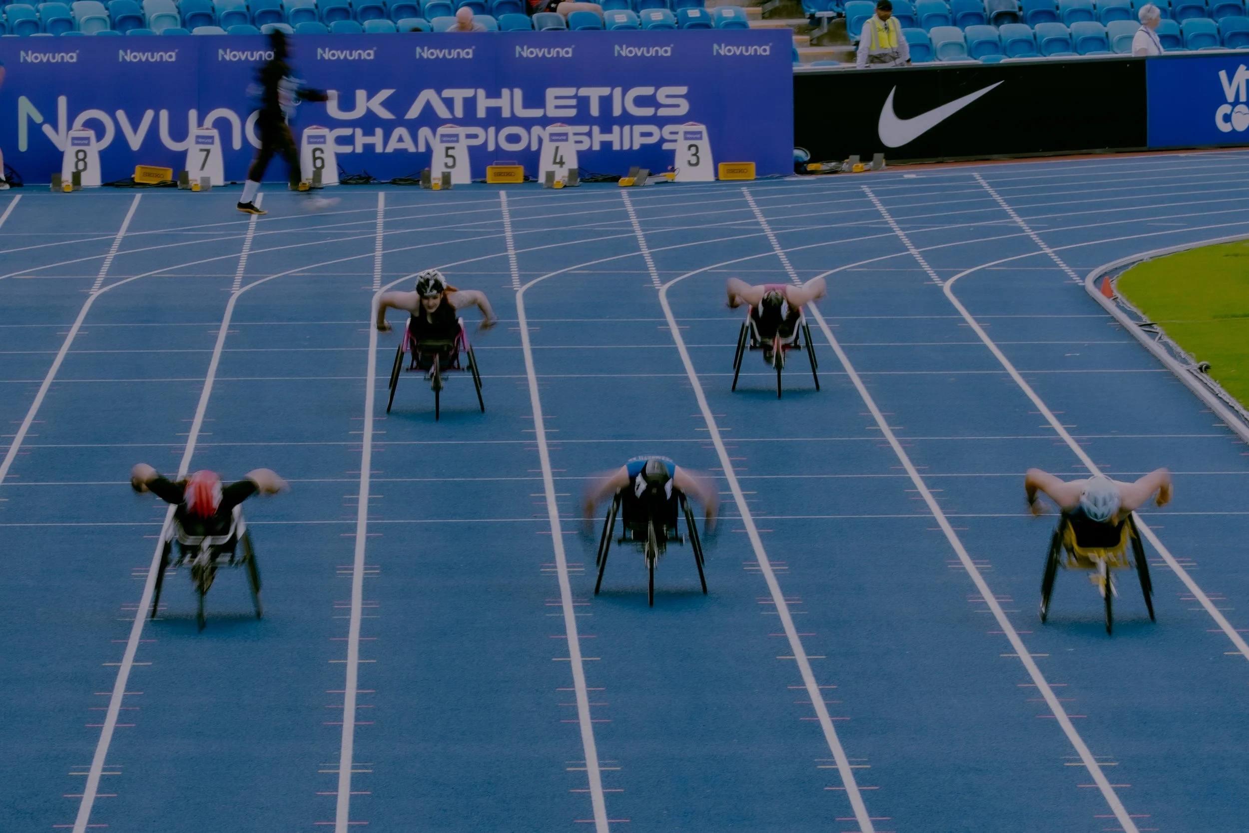 A wheelchair race at the UK Athletics Championships on a blue running track. Several athletes in wheelchairs are in position at the starting line, ready for the race. There are officials or staff on the sidelines, and a backdrop with the event name a