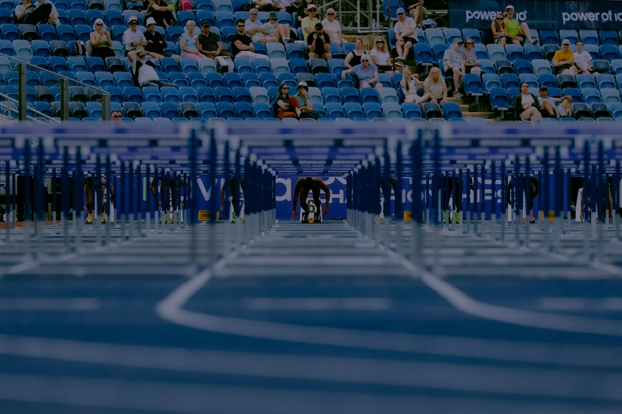 Crowd of spectators seated in stadium bleachers, watching an outdoor event, with some people taking photos and others wearing sunglasses and hats.