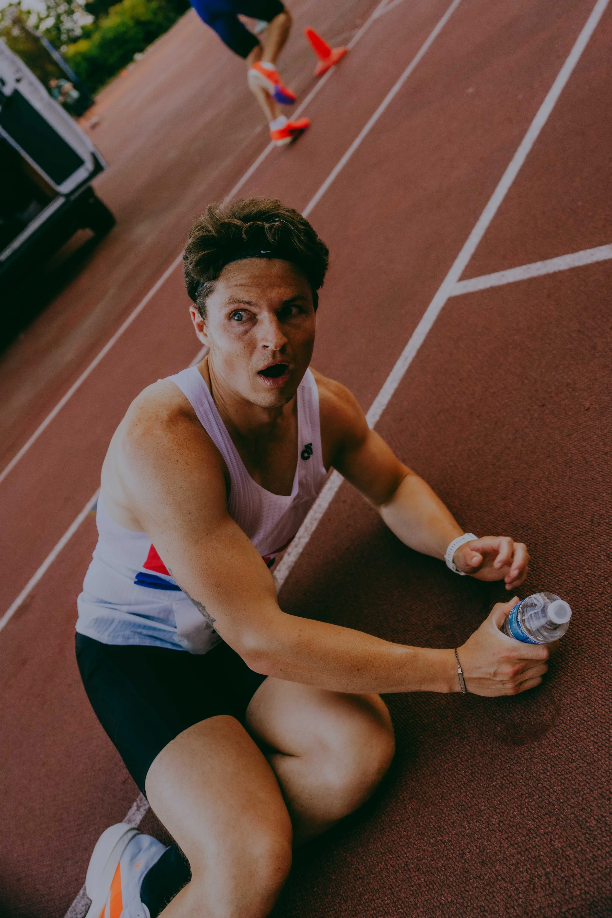 A young man in athletic clothing sitting on a running track with a water bottle in his hand and an exhausted expression, having just finished a race.