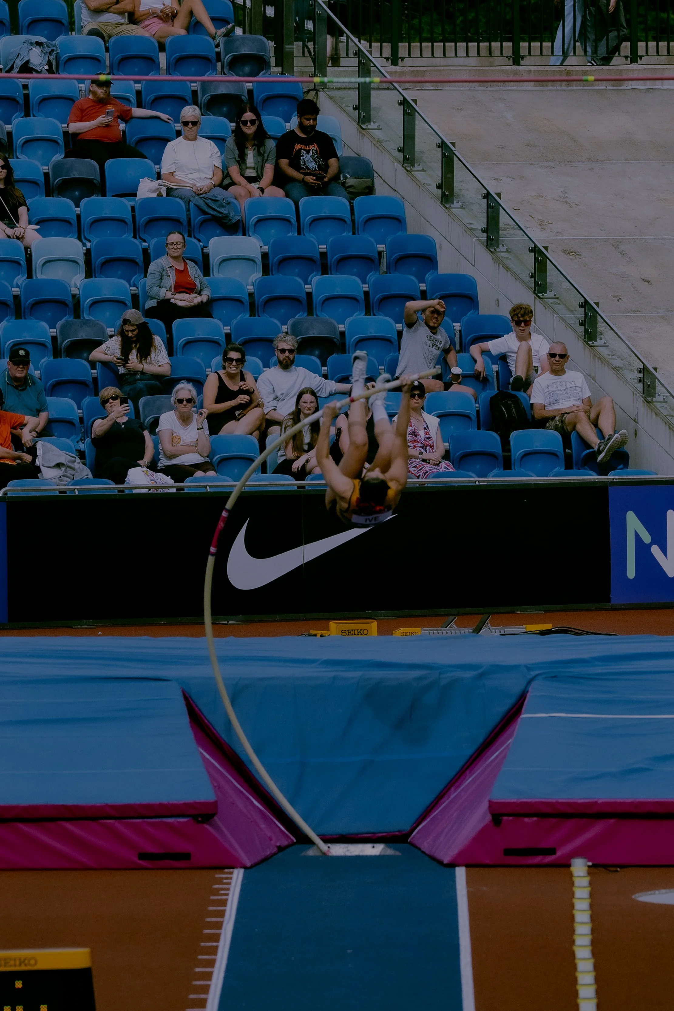 An athlete in mid-flight during a pole vault attempt over a pink and blue bar at a track and field event, with spectators seated in the background.