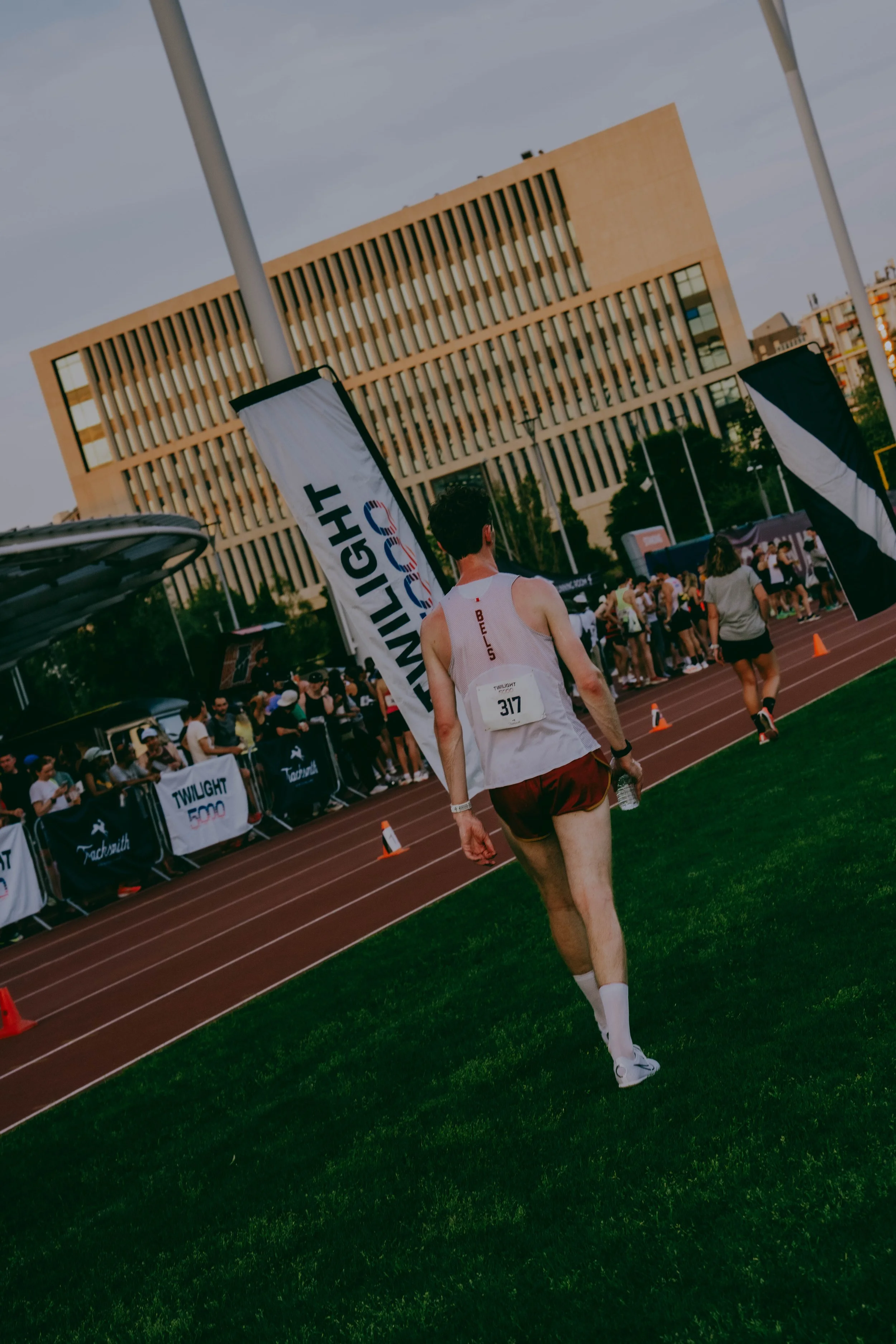 A male runner wearing a white tank top and maroon shorts at a track meet during the daytime, with a large building and a crowd of spectators in the background.