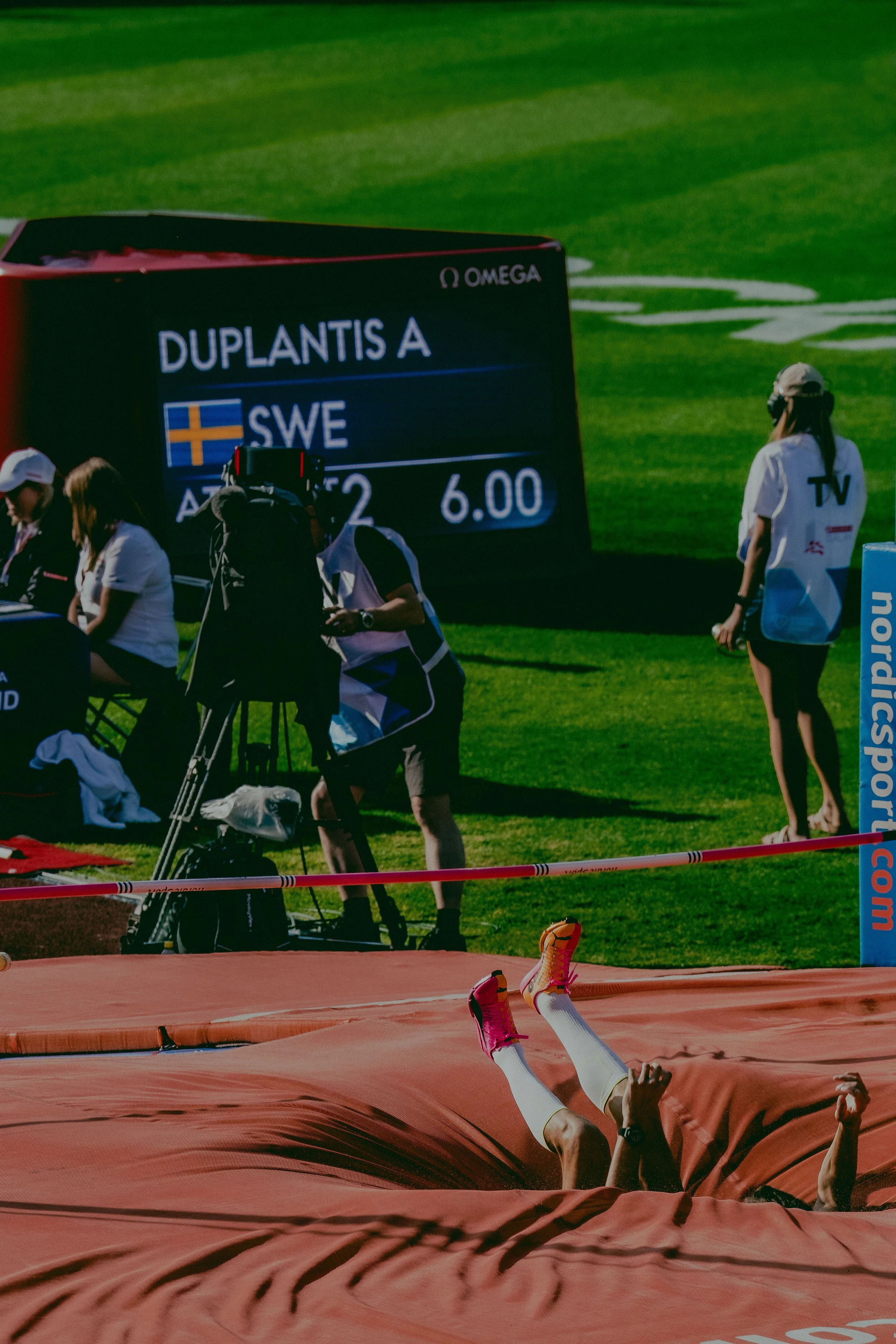 Track and field stadium with fallen athlete at the pole vault event, surrounded by officials and camera crew, scoreboard displaying athlete names and scores, green grass field in the background.