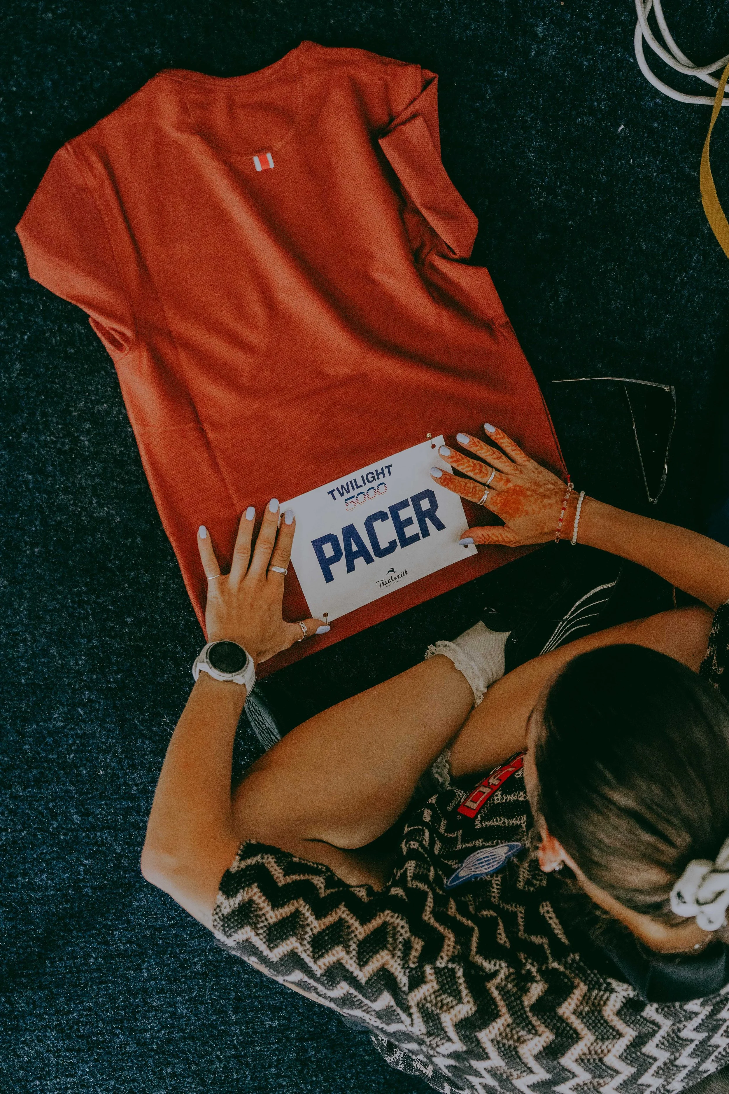 A person with a scanner on their wrist, wearing a black and white zigzag patterned shirt and white socks, is sitting on dark blue carpeted floor. They are holding a race bib labeled 'TWILIGHT 5000 RACER' with both hands, one hand over the bib, and th