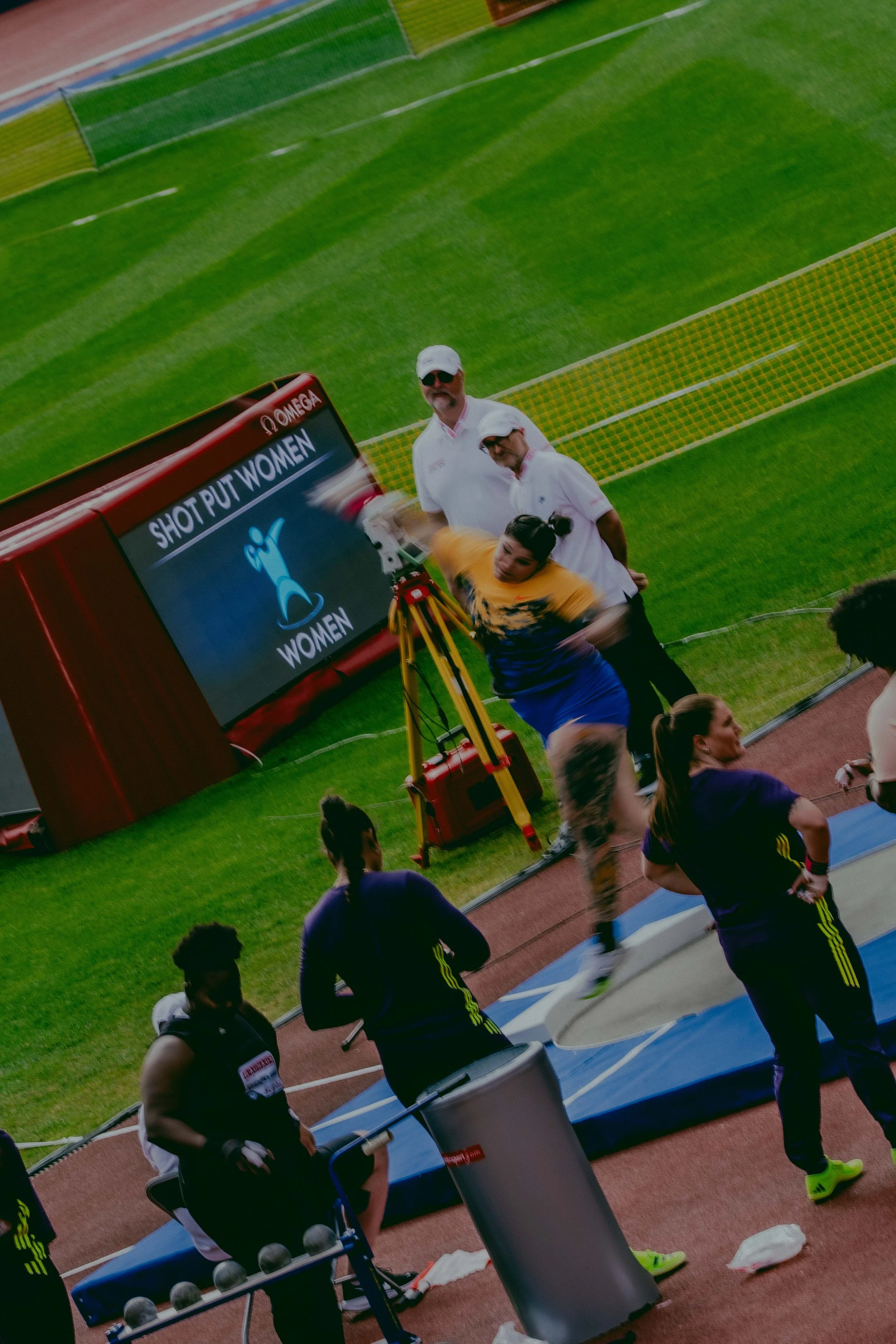 People running on a track during a women's athletic event. Several athletes are visible with some wearing athletic gear and others standing nearby, with a green field and stadium seating in the background.