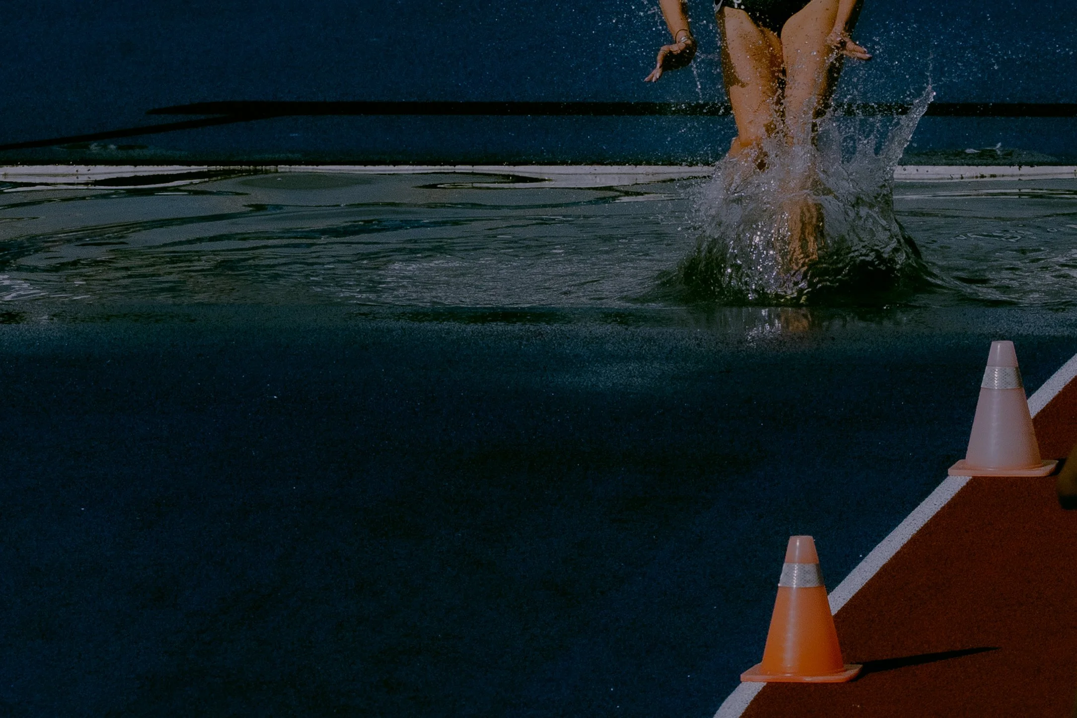 An athlete is diving into a water pool during a race, with a traffic cone placed on the starting line to the right.