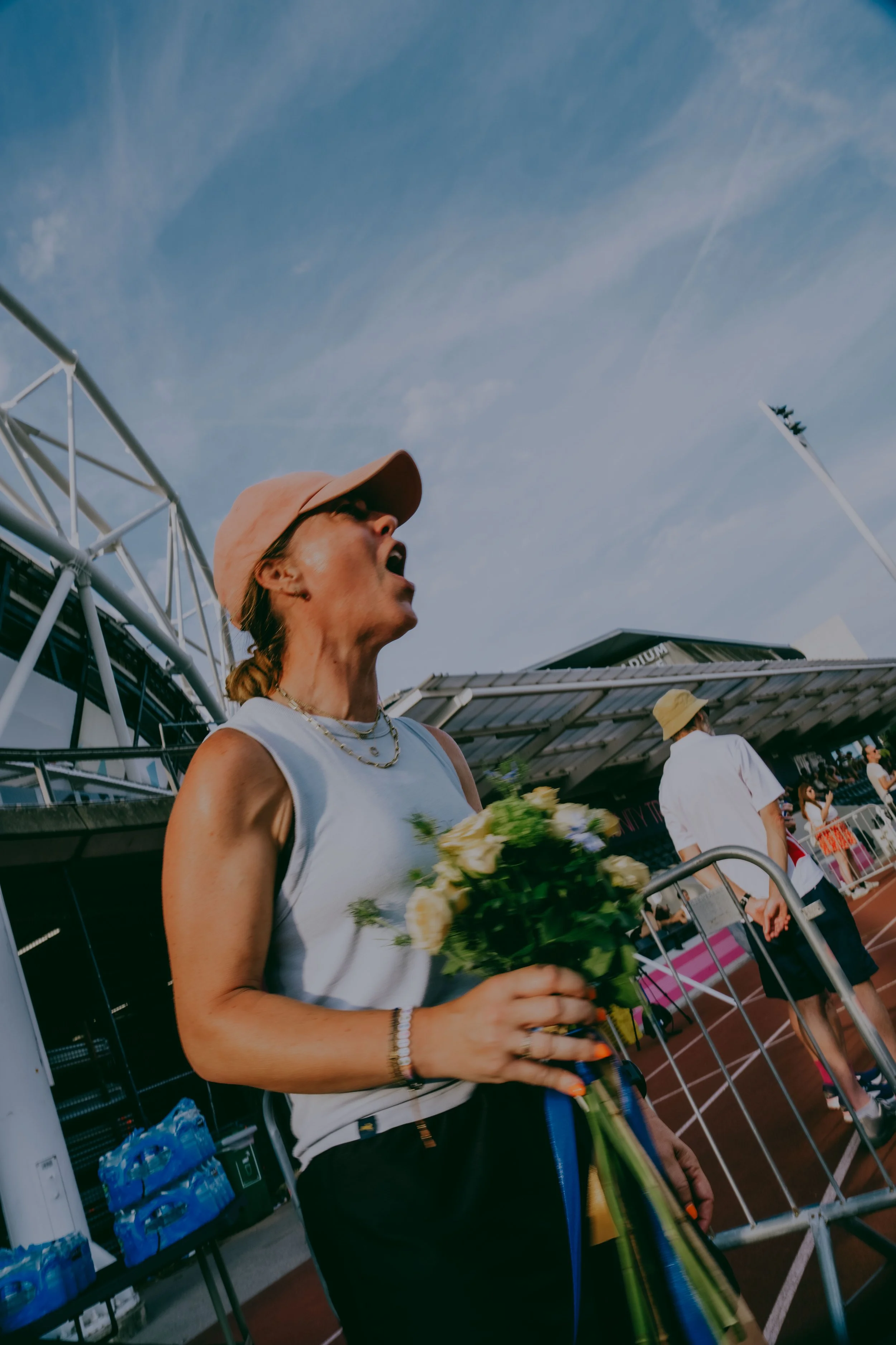 A woman wearing a tan baseball cap, white sleeveless top, and gold jewelry holding a bouquet of yellow and white flowers, standing in front of a stadium entrance with people entering behind her.