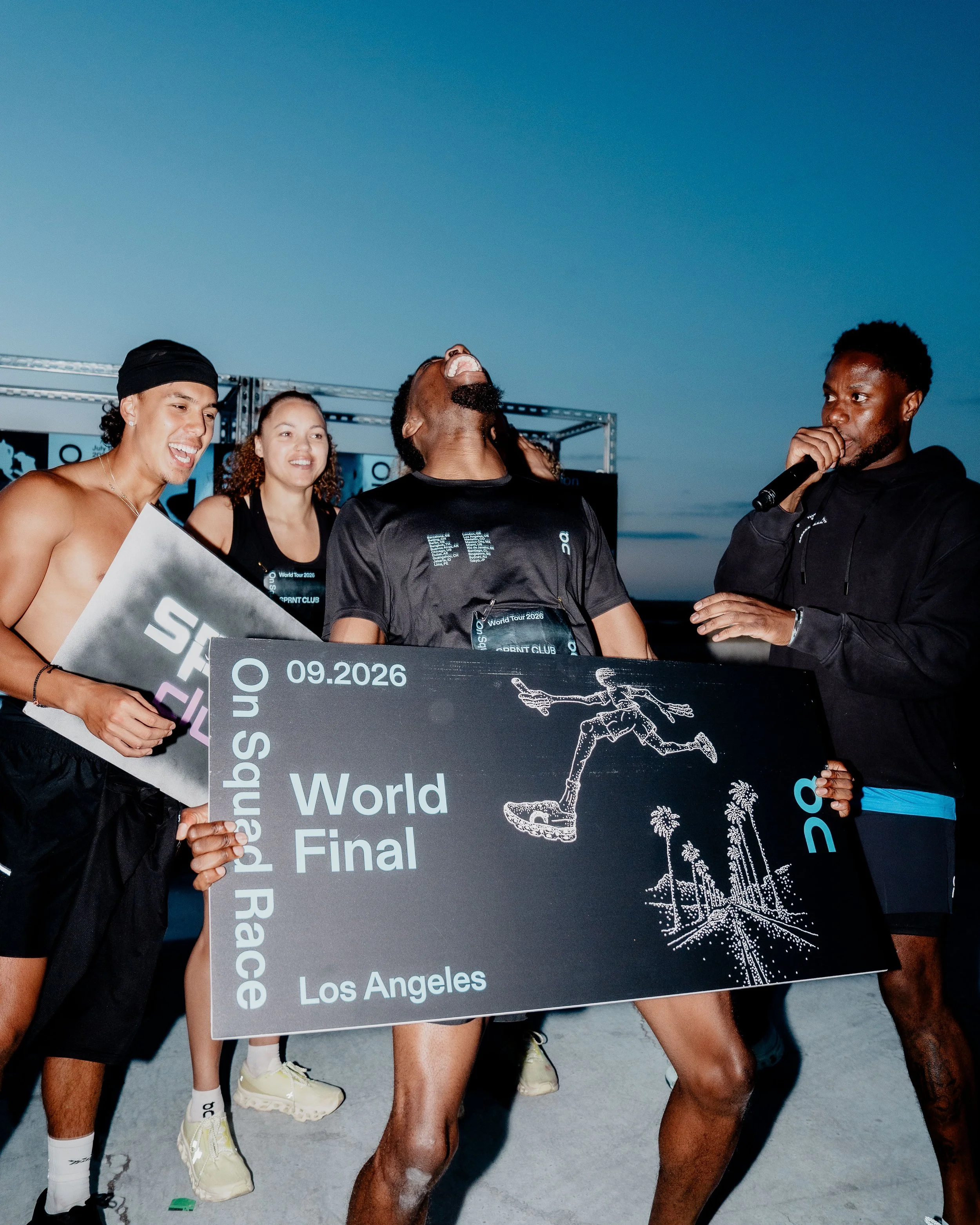 Group of runners at the Los Angeles batona race, with one runner holding a sign that reads 'World Tour 2026 Los Angeles' and they are dressed in athletic clothing, celebrating after finishing a race.