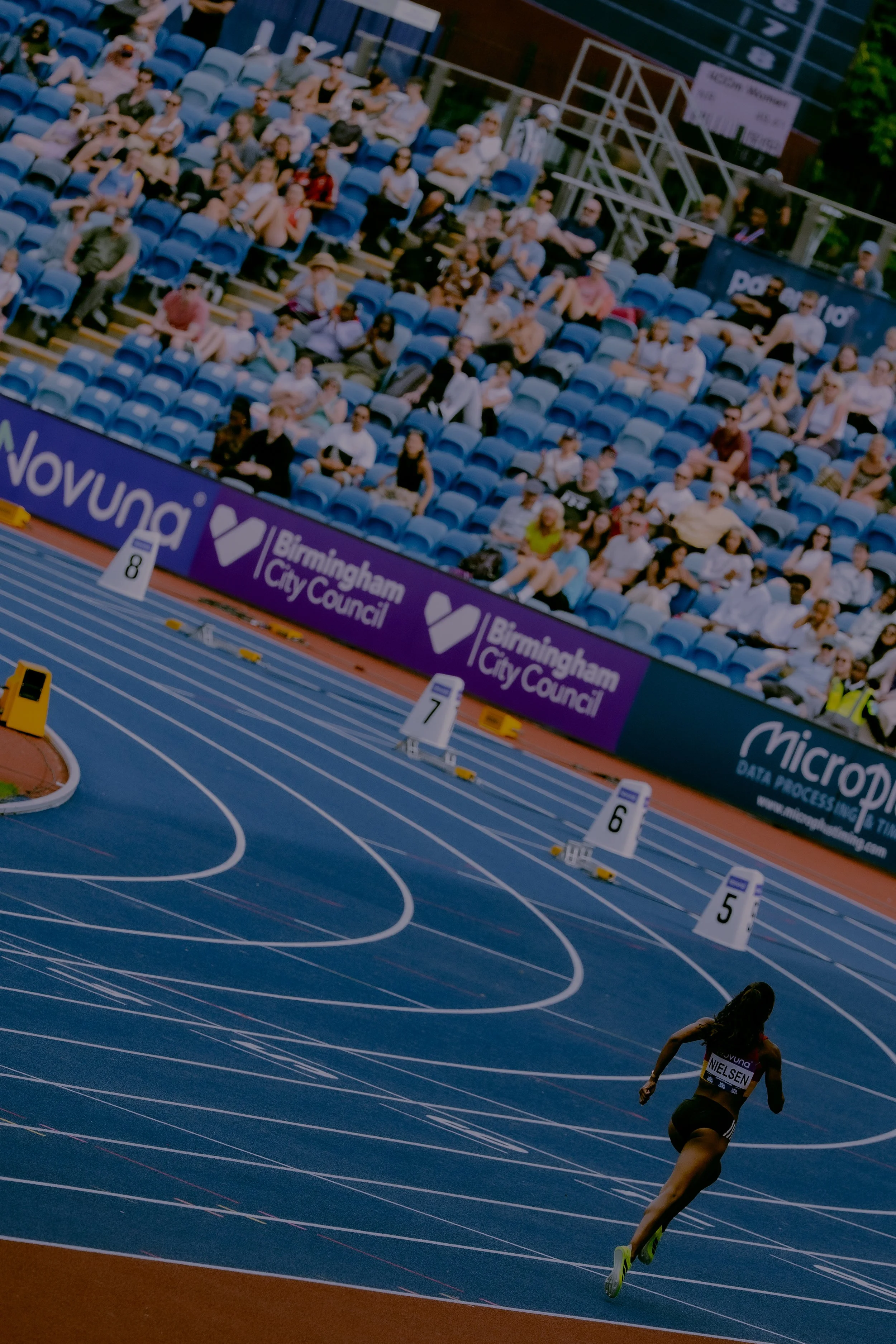 A female athlete sprinting on a blue indoor track with numbered lanes, near a crowd sitting in blue stadium seats, with a Birmingham City Council banner in the background.