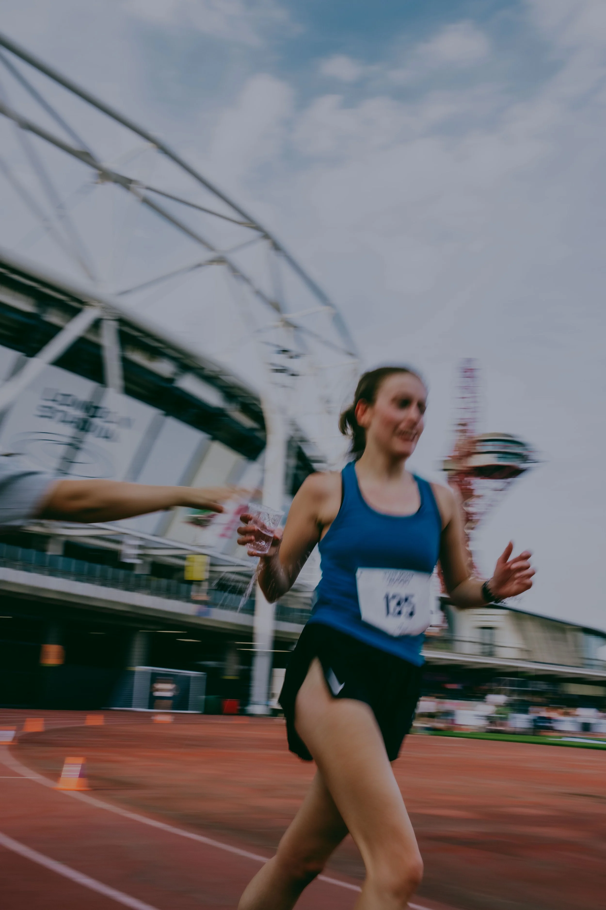 A female runner wearing a blue top and black shorts, holding a water cup while running on a track during daytime, with a modern stadium and cloudy sky visible in the background.