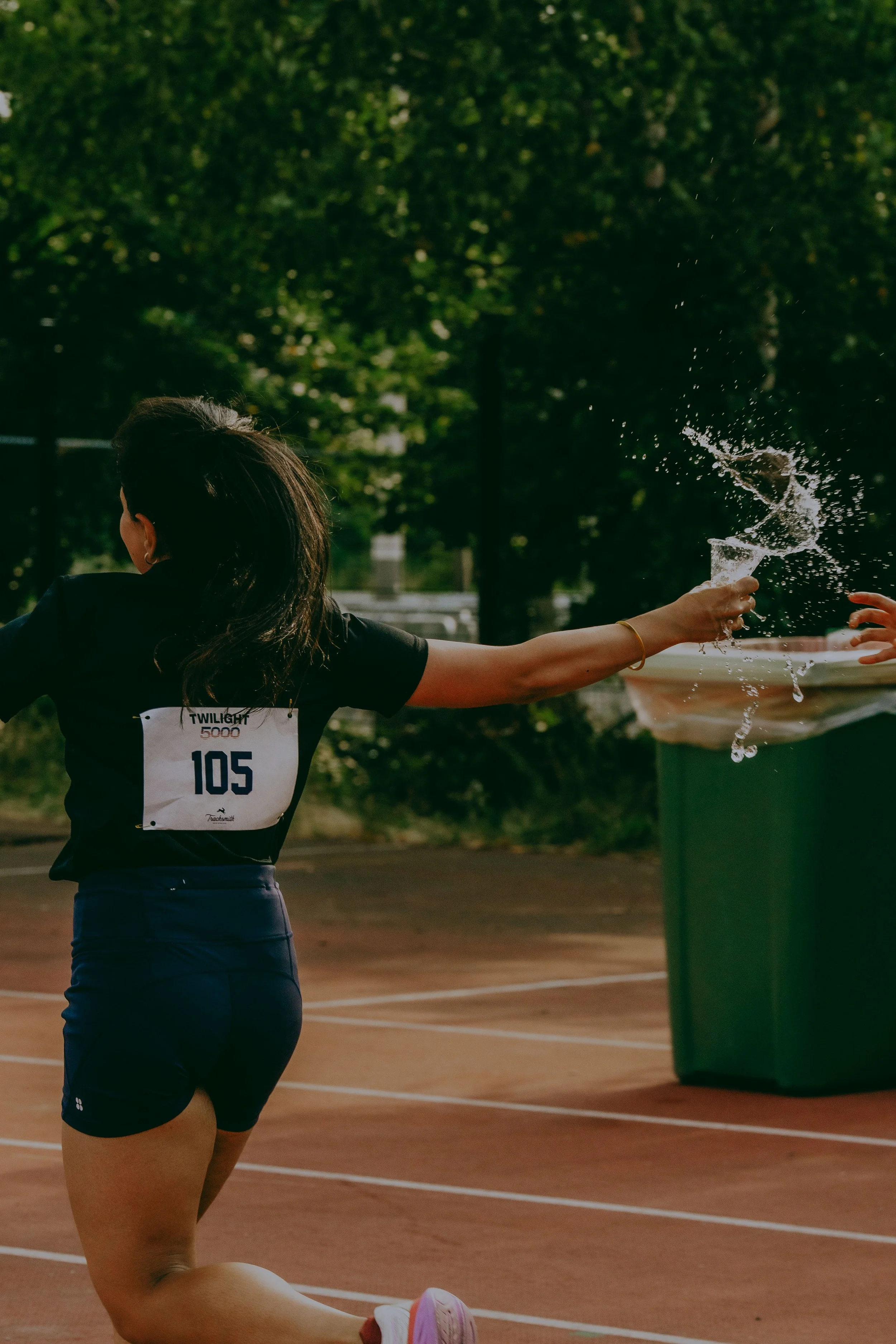 A woman participating in a running event on a track, holding a water cup and splashing water on herself, with trees in the background.