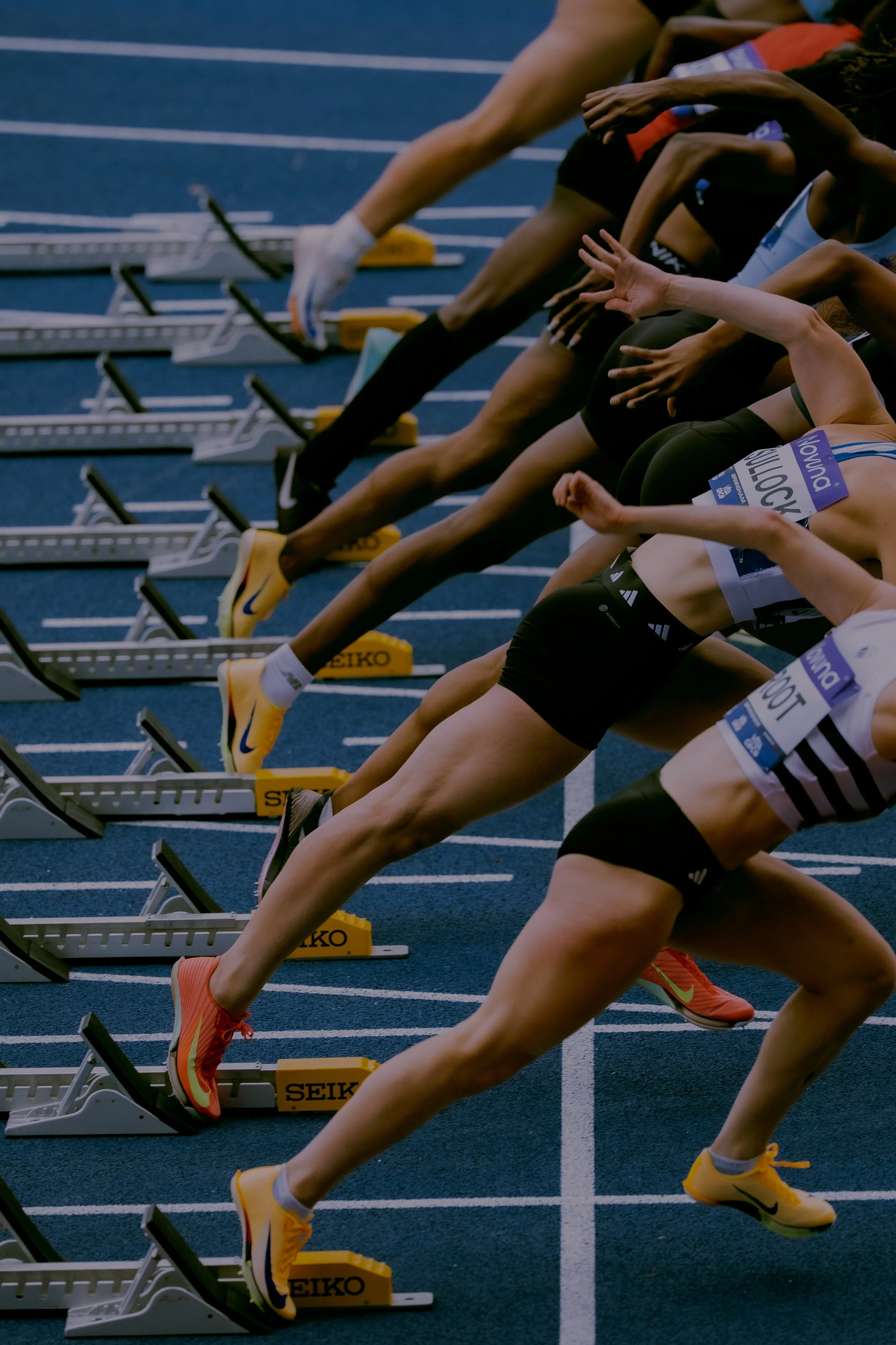 Multiple athletes at the starting line for a track race, crouched in starting positions on a blue running track with hurdles labeled 'Seiko' in the foreground.