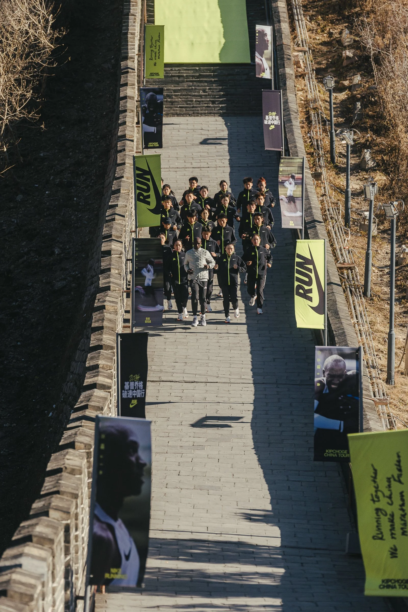 A group of people dressed in black athletic jackets walking down concrete stairs outdoors. There are flags with Nike branding and Chinese characters, some featuring images of athletes. The background includes leafless trees and rocky terrain.