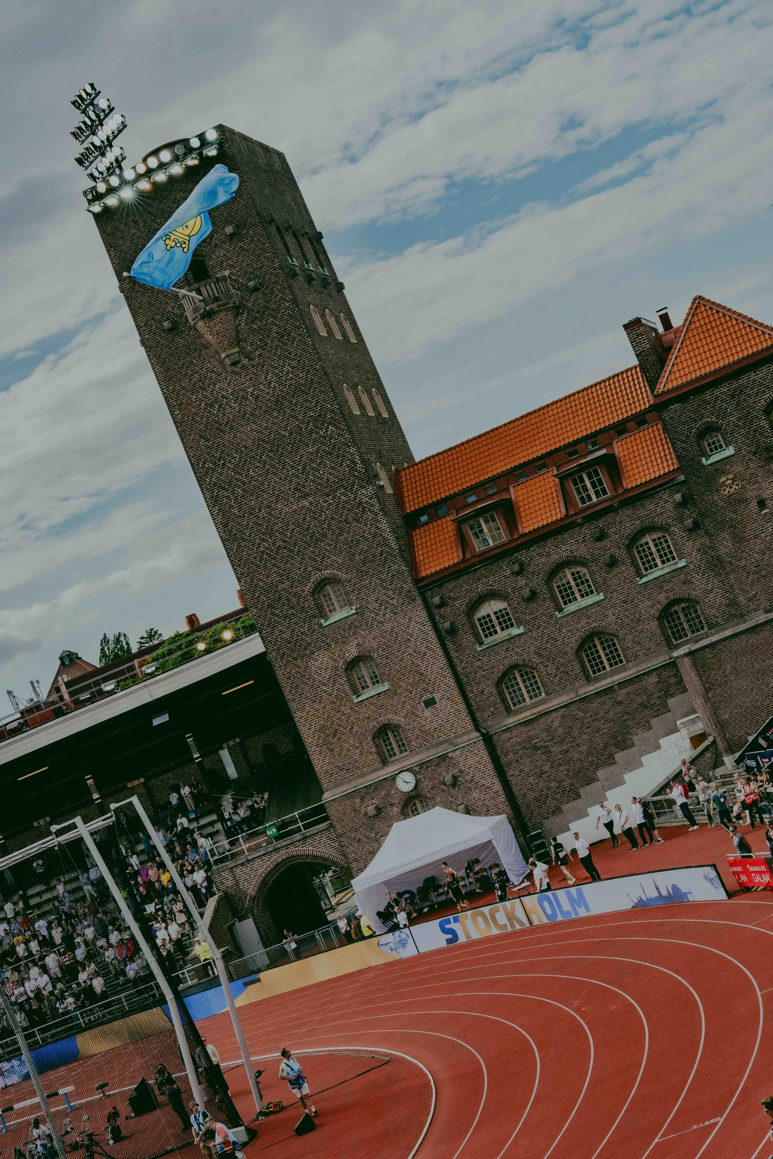 A view of a large brick athletic stadium with a tall tower featuring bright lights and a Swedish flag. The stadium has a red running track with multiple lanes, a section with spectators, and a white tent at the edge. The sky is partly cloudy, and the