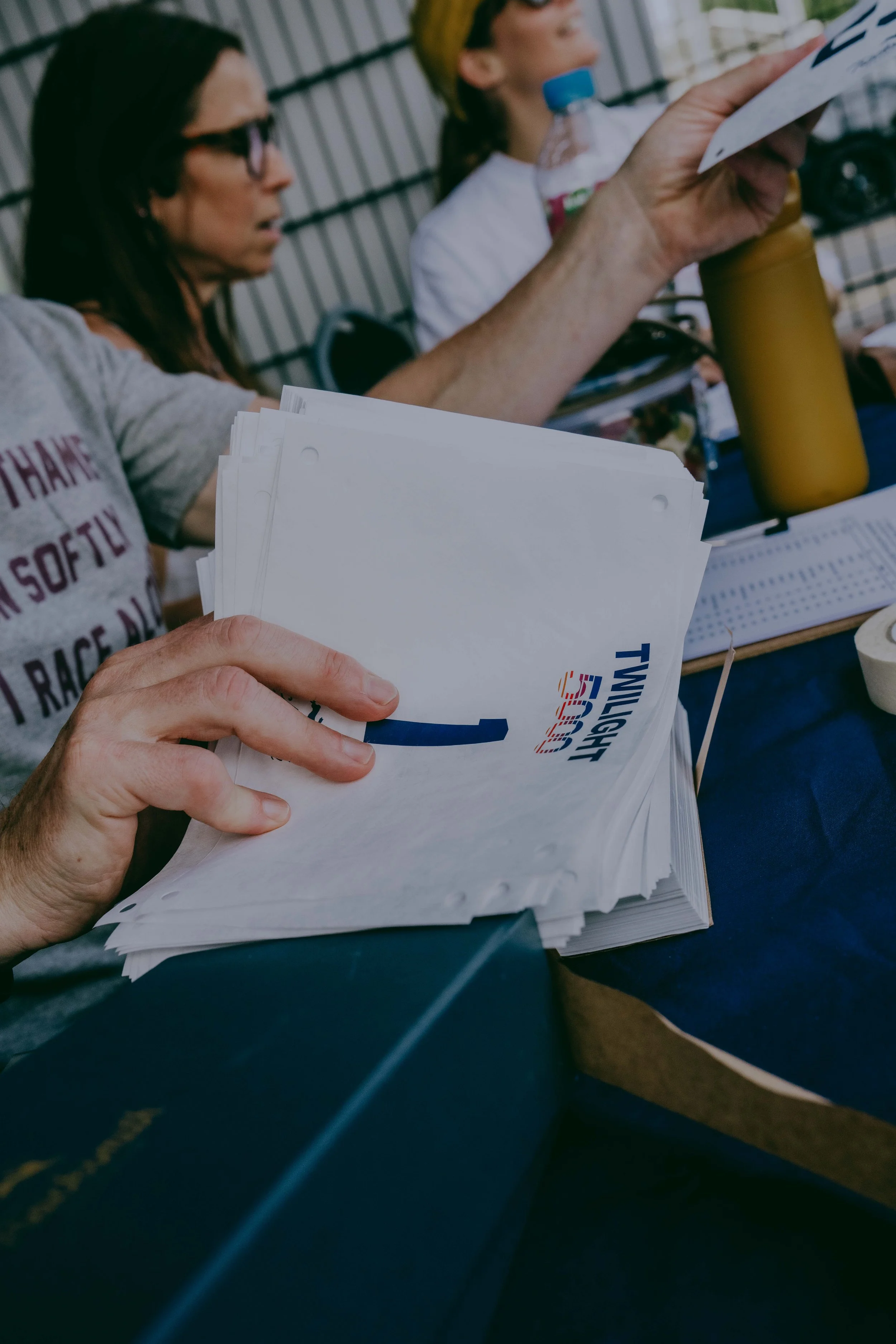 A person holding a stack of printed papers, with a woman and a child sitting at a table in the background. The woman is looking down, and the child is drinking from a bottle. The table has a yellow water bottle, a document, and a pen.