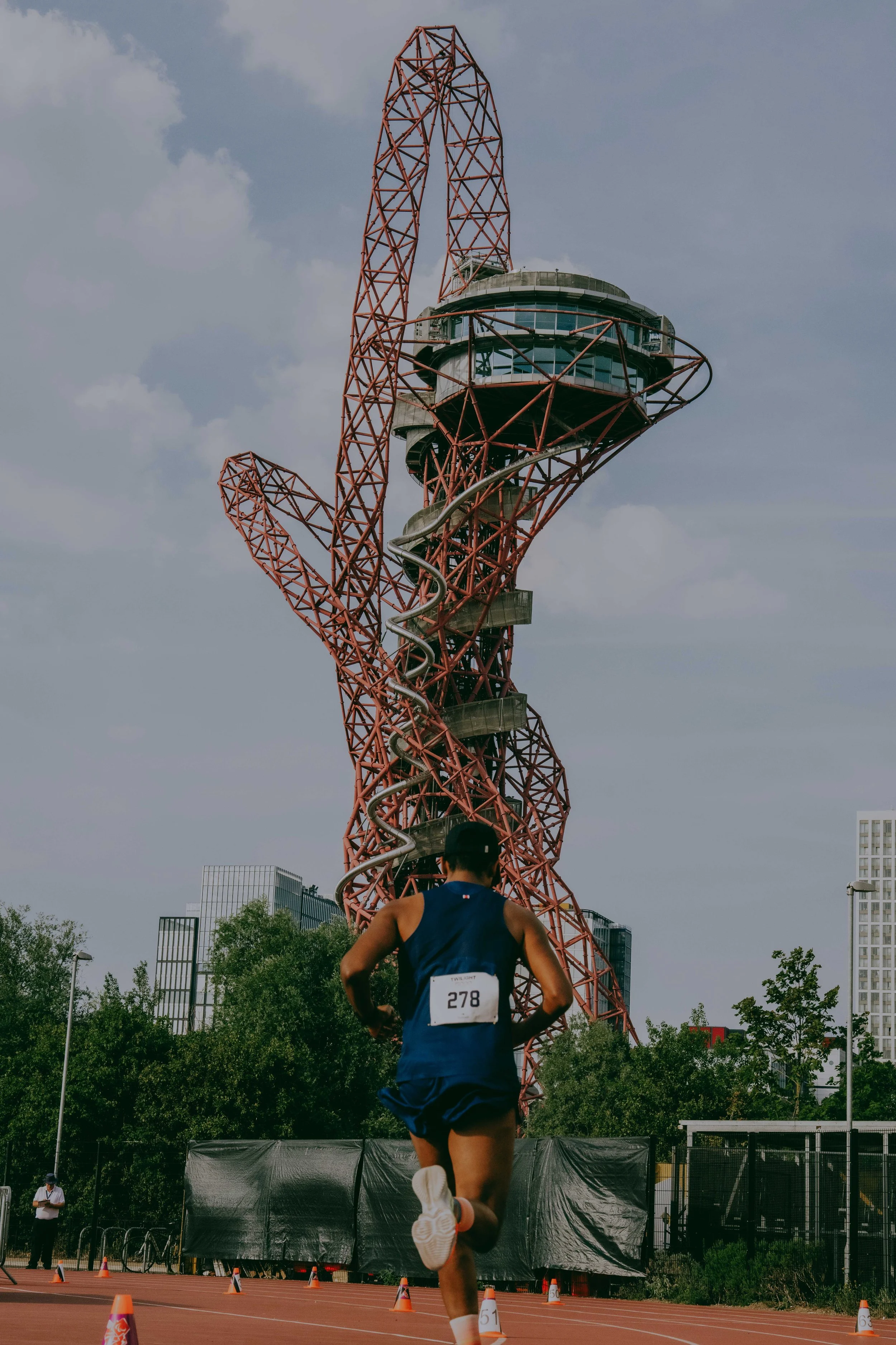 A marathon runner wearing a blue uniform with bib number 278 runs on a track with orange cones. In the background, there is a tall, red, lattice-like tower with a spiral slide and an observation deck, with several high-rise buildings and trees around