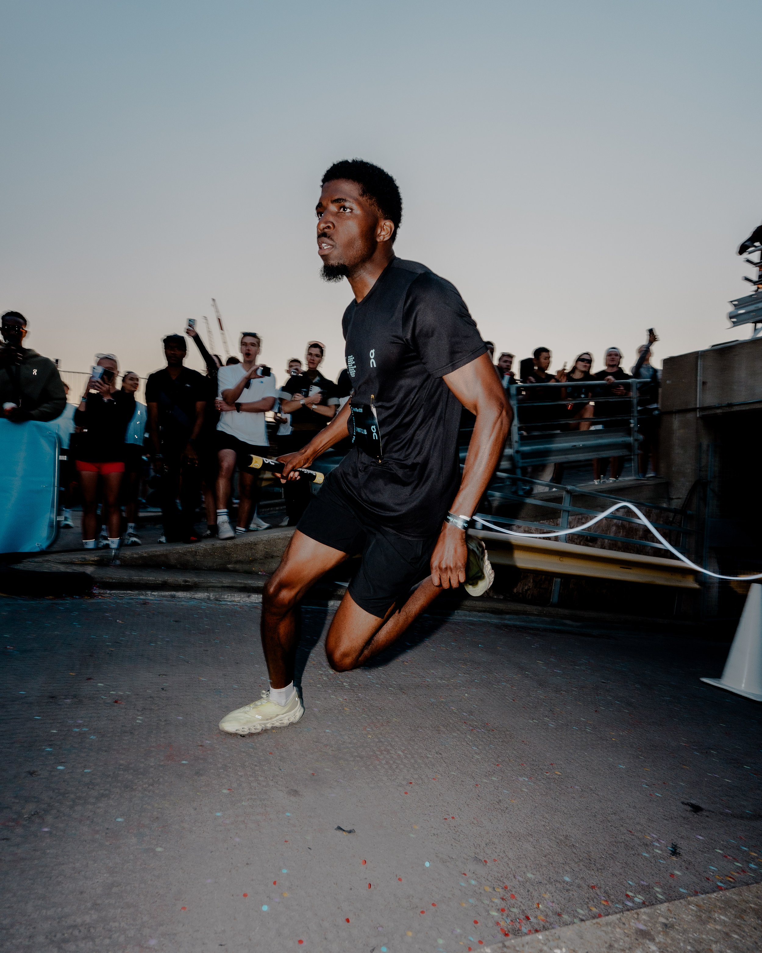 A man in black athletic clothing kneels on the ground with a group of people in the background. The man is holding a medal, and there are colorful confetti on the ground. It appears to be an outdoor event taking place at dusk or evening.