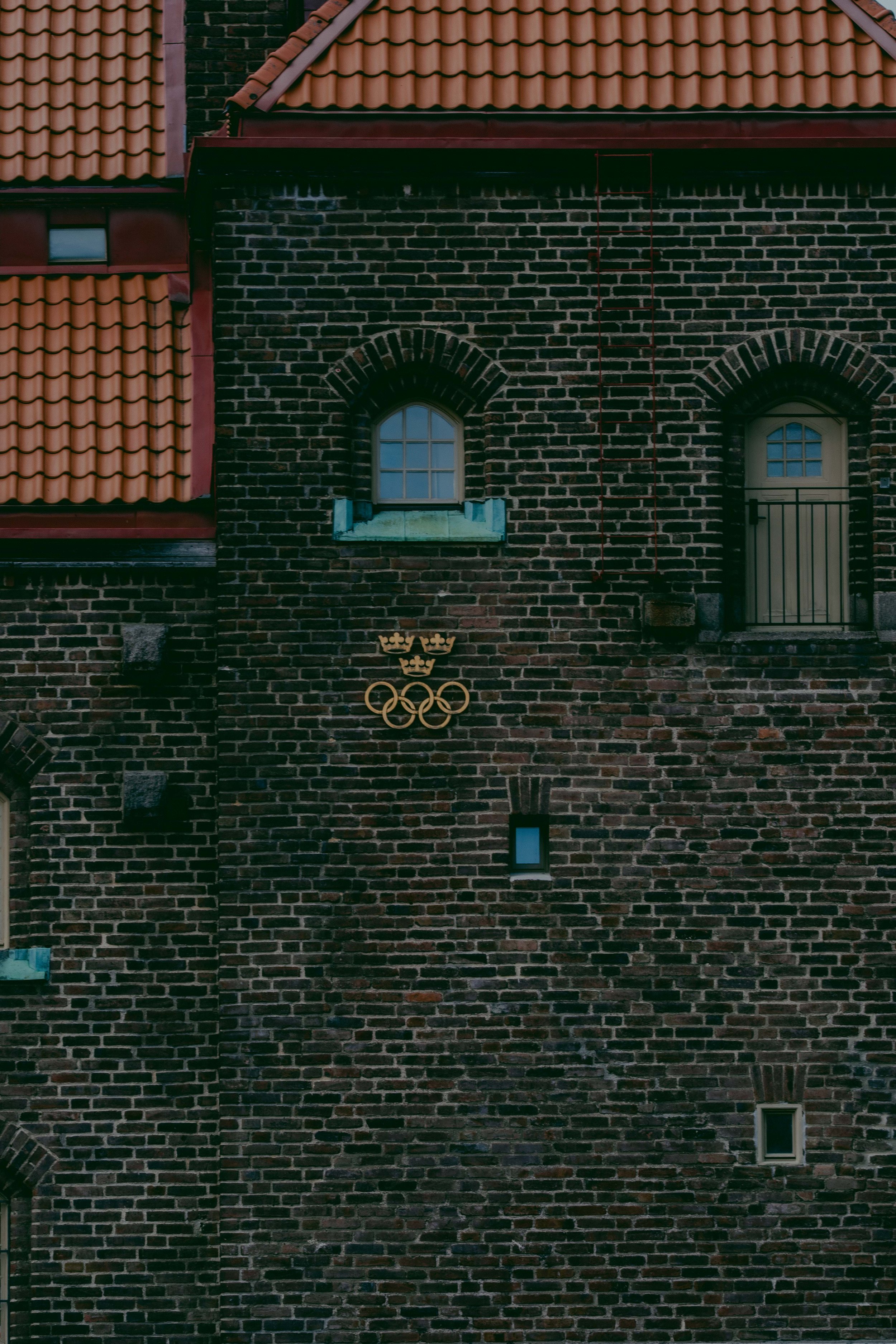 Close-up of a brick building wall with three small windows, a red ladder, and the Olympic rings symbol with three crown icons below.