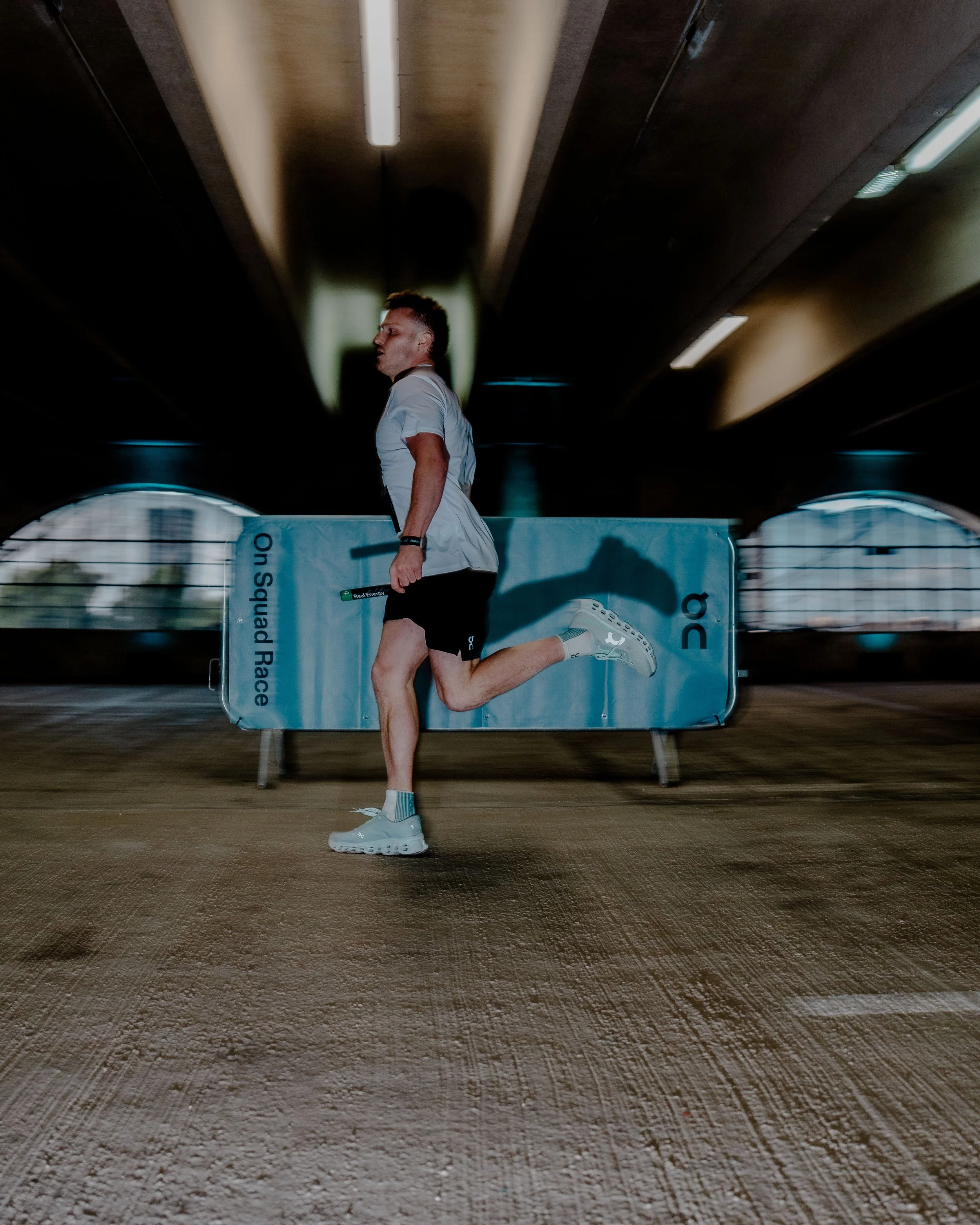 A person running in an indoor sports arena with a dark ceiling and bright lights. The runner is wearing a white shirt, black shorts, and white running shoes. A banner behind them displays some text, and the floor is a textured surface.