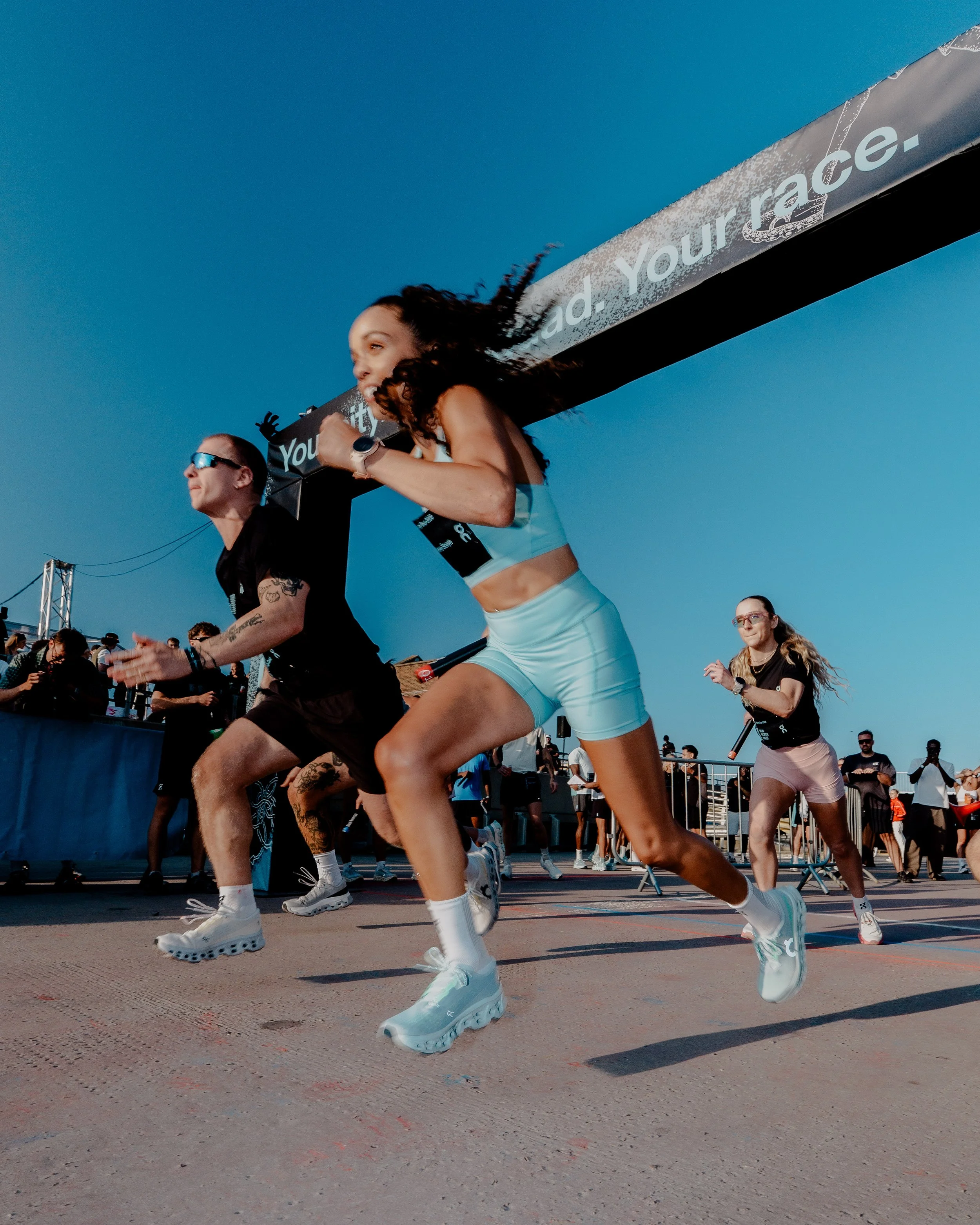 People running in a race during a sports event on a sunny day, with a large banner overhead and a crowd in the background.