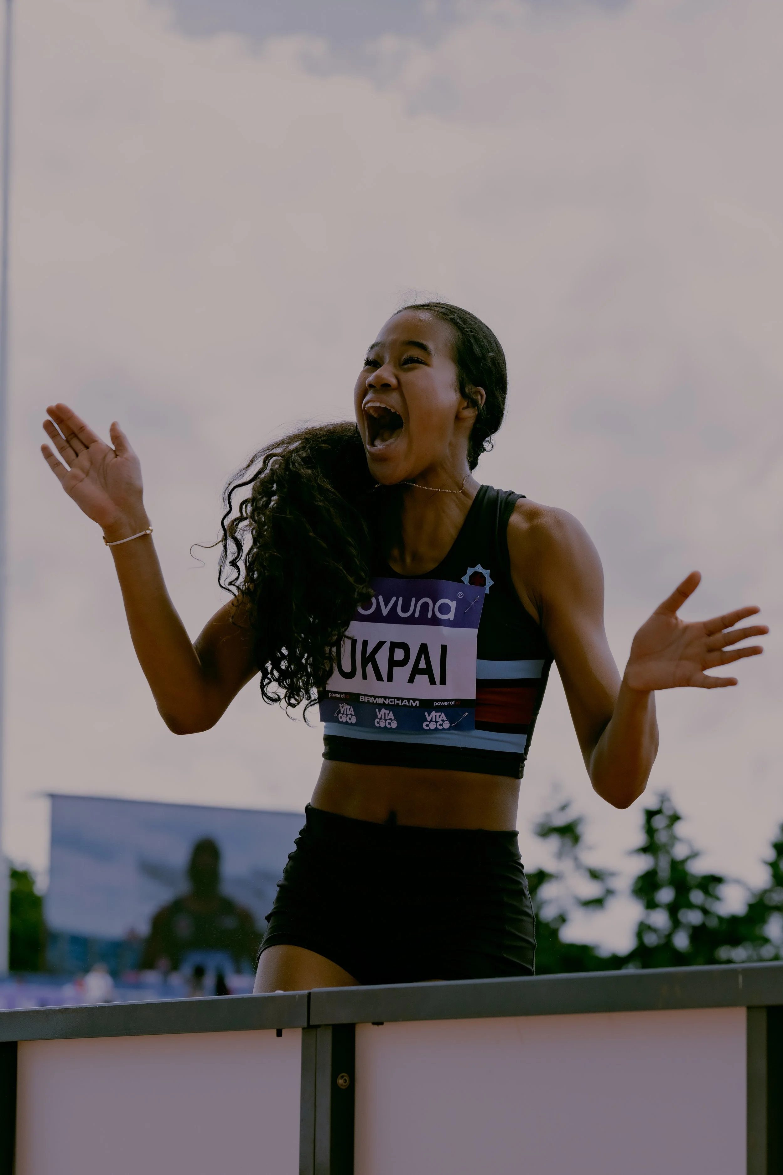 Young woman wearing athletic clothes celebrating at a sports event, with her arms raised and facial expression showing excitement.