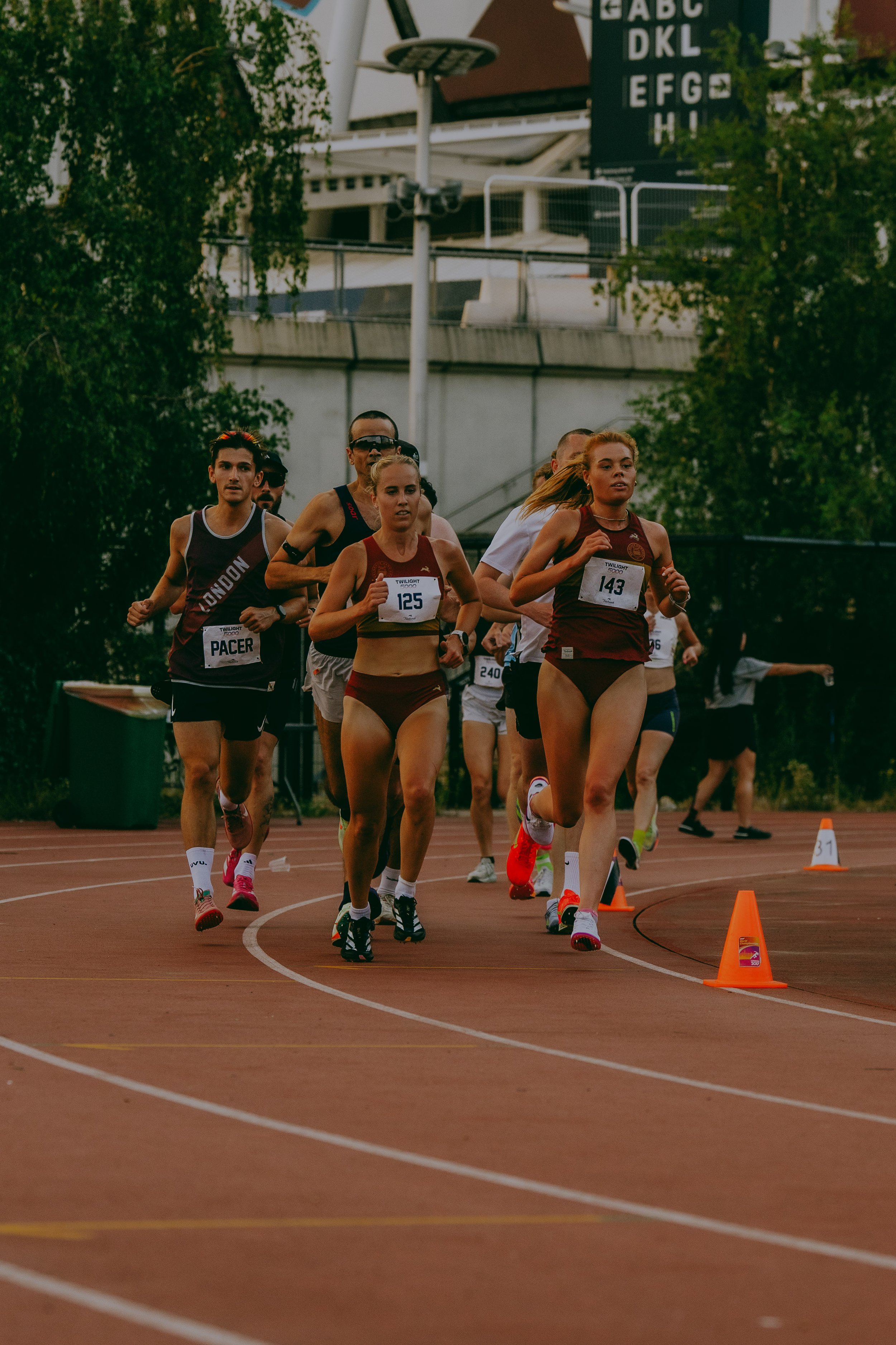 A group of runners participating in a track race, running on a red track with orange cones marking the course. The runners are wearing athletic clothing and race numbers, with a background of trees, a cityscape, and an overhead sign with letters.