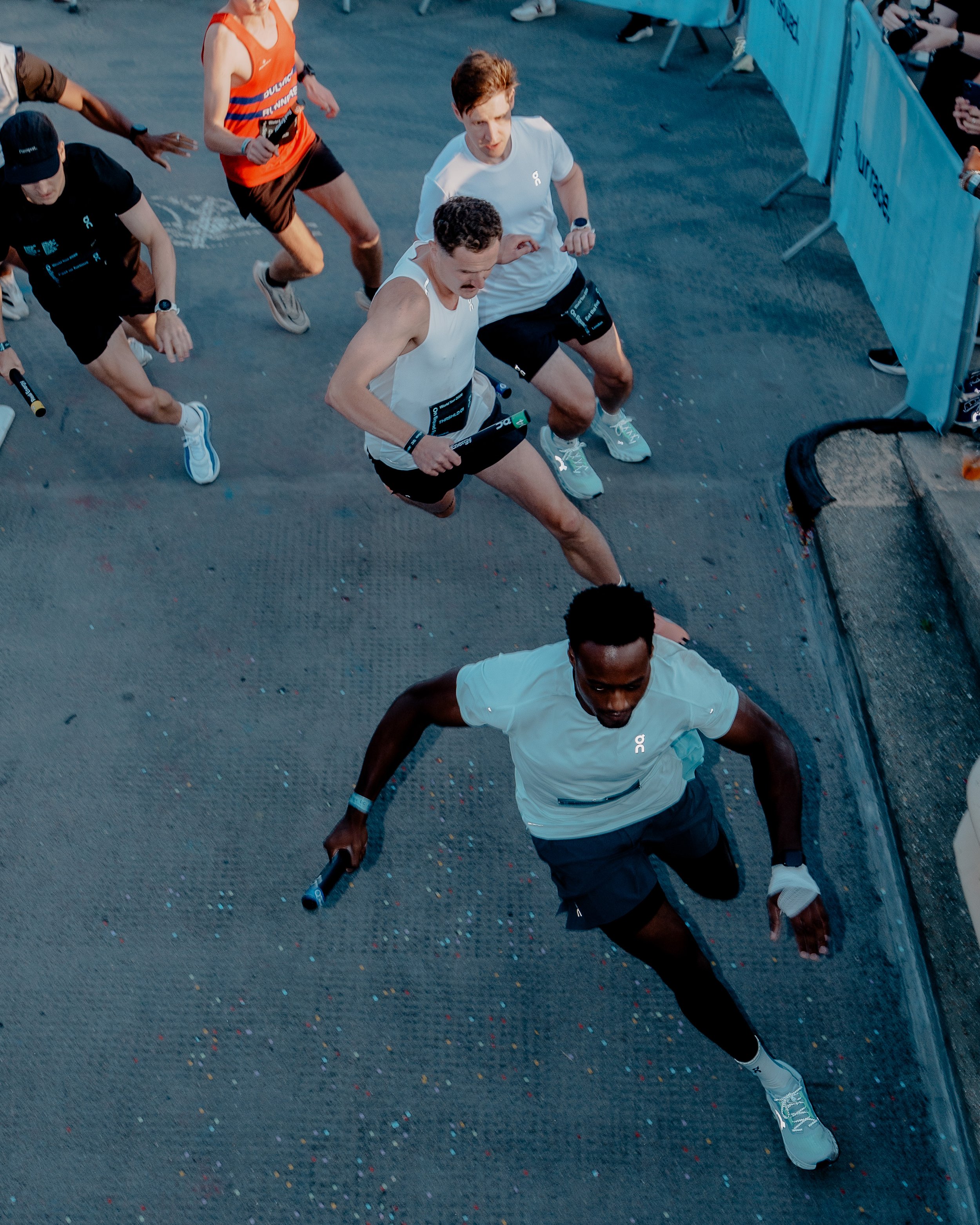 Group of runners in a race, wearing athletic gear and running on a track with colorful confetti on the ground.