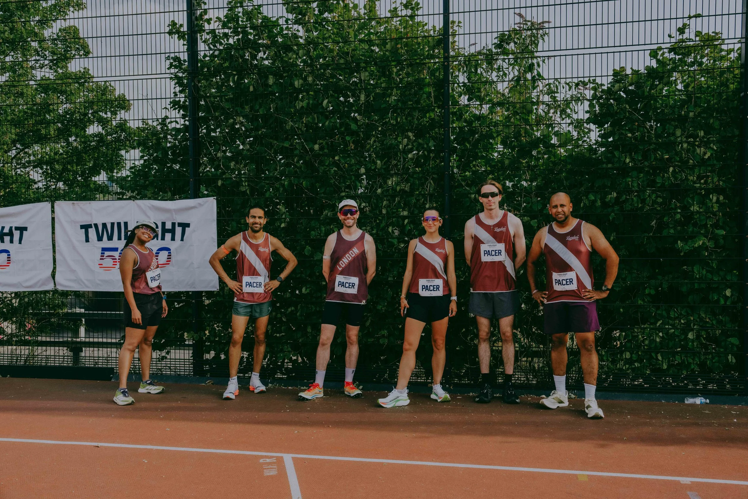 Group of six runners in athletic gear standing on a tennis court in front of a tall fence with green trees behind it, posing for a photo before a race.
