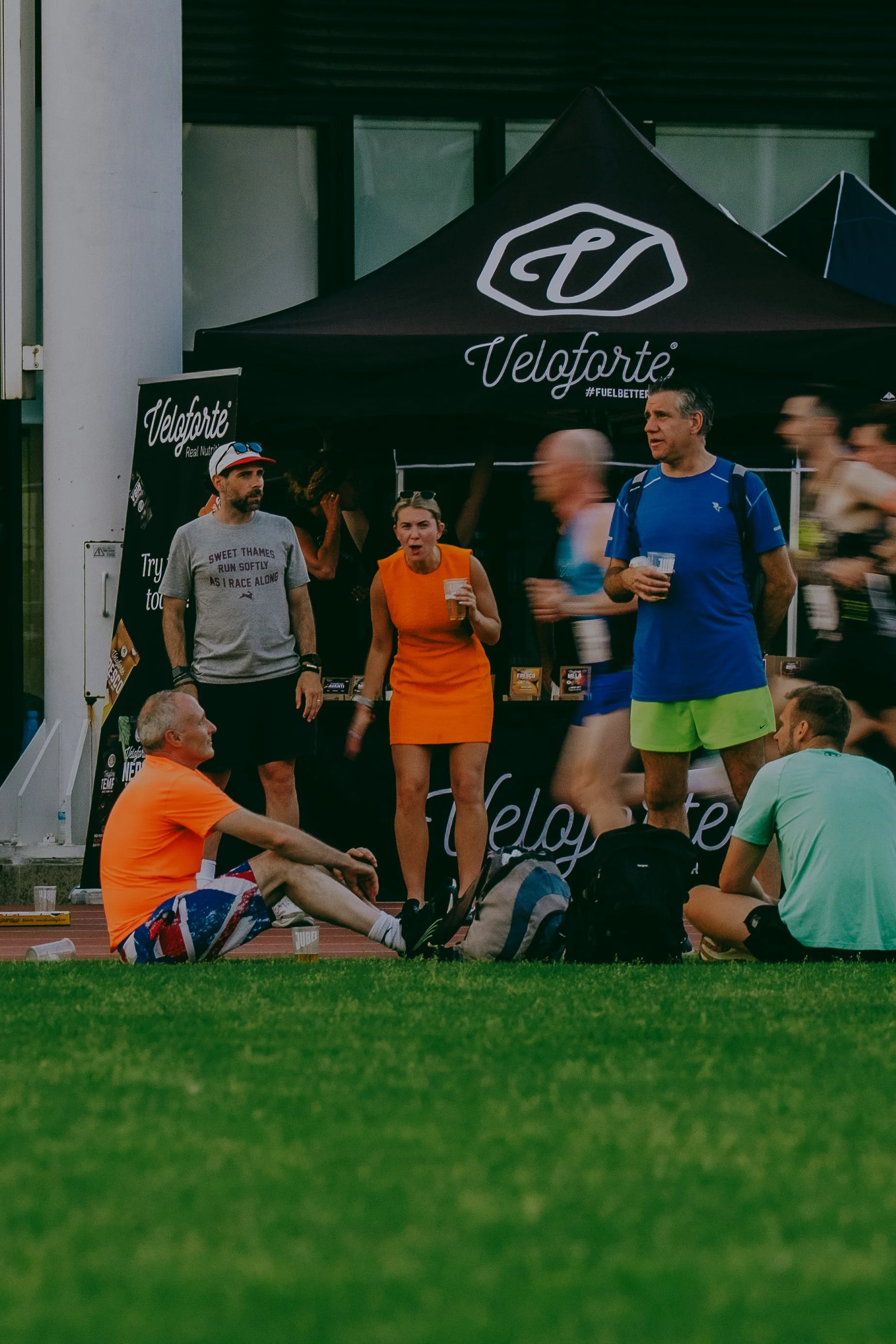 Group of people resting and talking at an outdoor sports event, with a black tent branded 'Velo forte' in the background. Some are sitting on the ground, while others are standing.