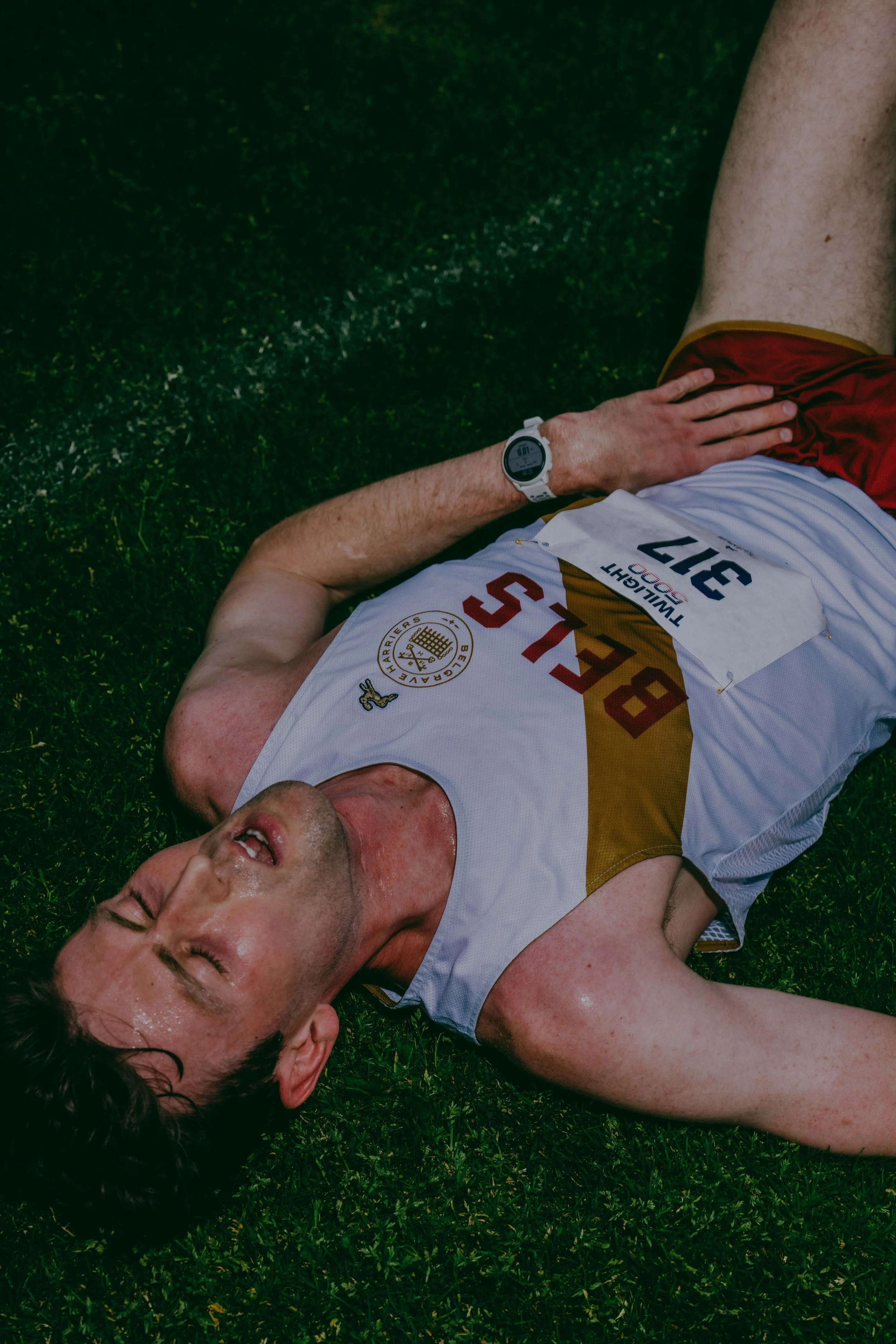 A man lying on grass, appearing to be exhausted or hurt, wearing a white sports jersey with red and gold accents, and a white wristwatch.