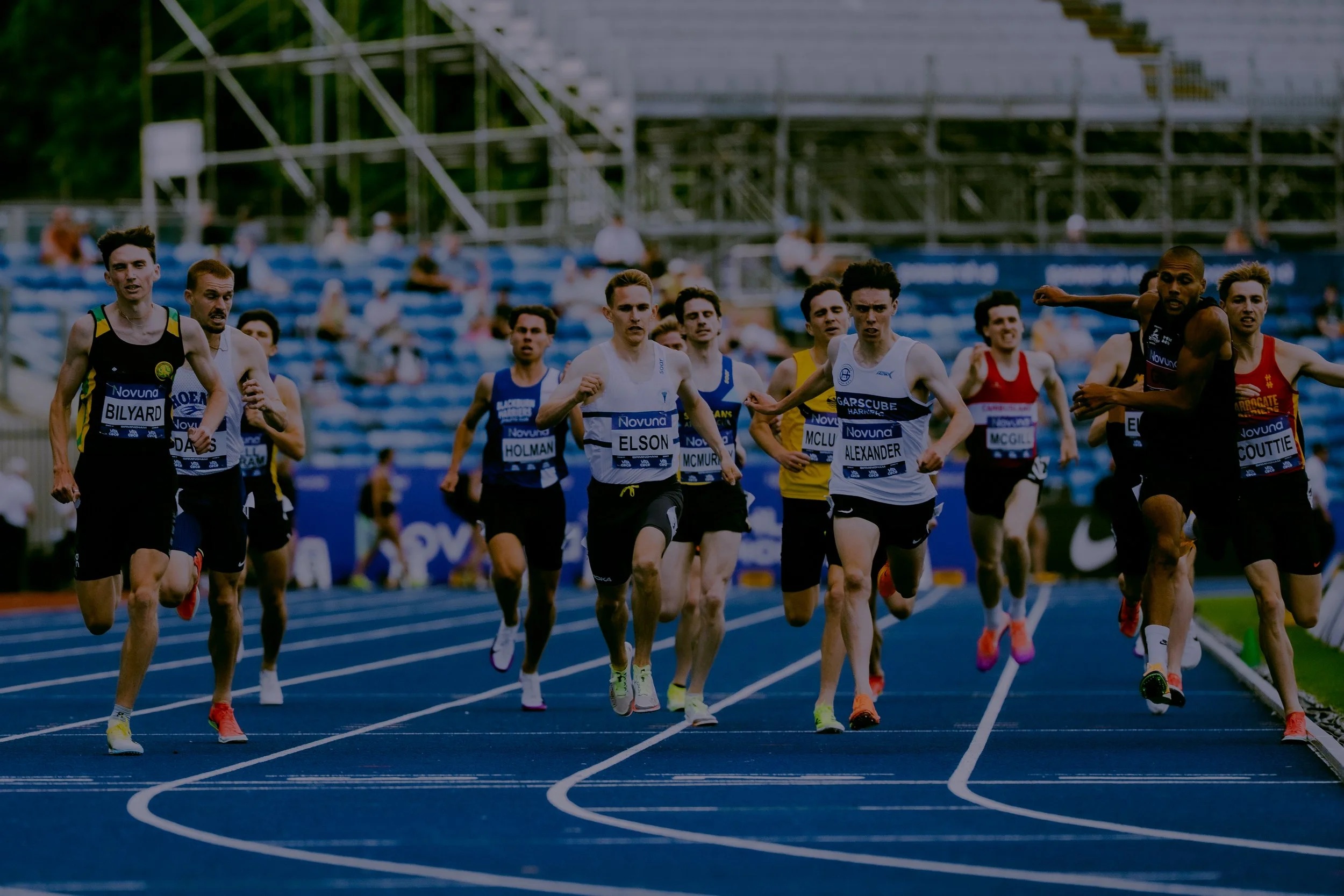 A group of male athletes running on a blue track in a stadium during a race.